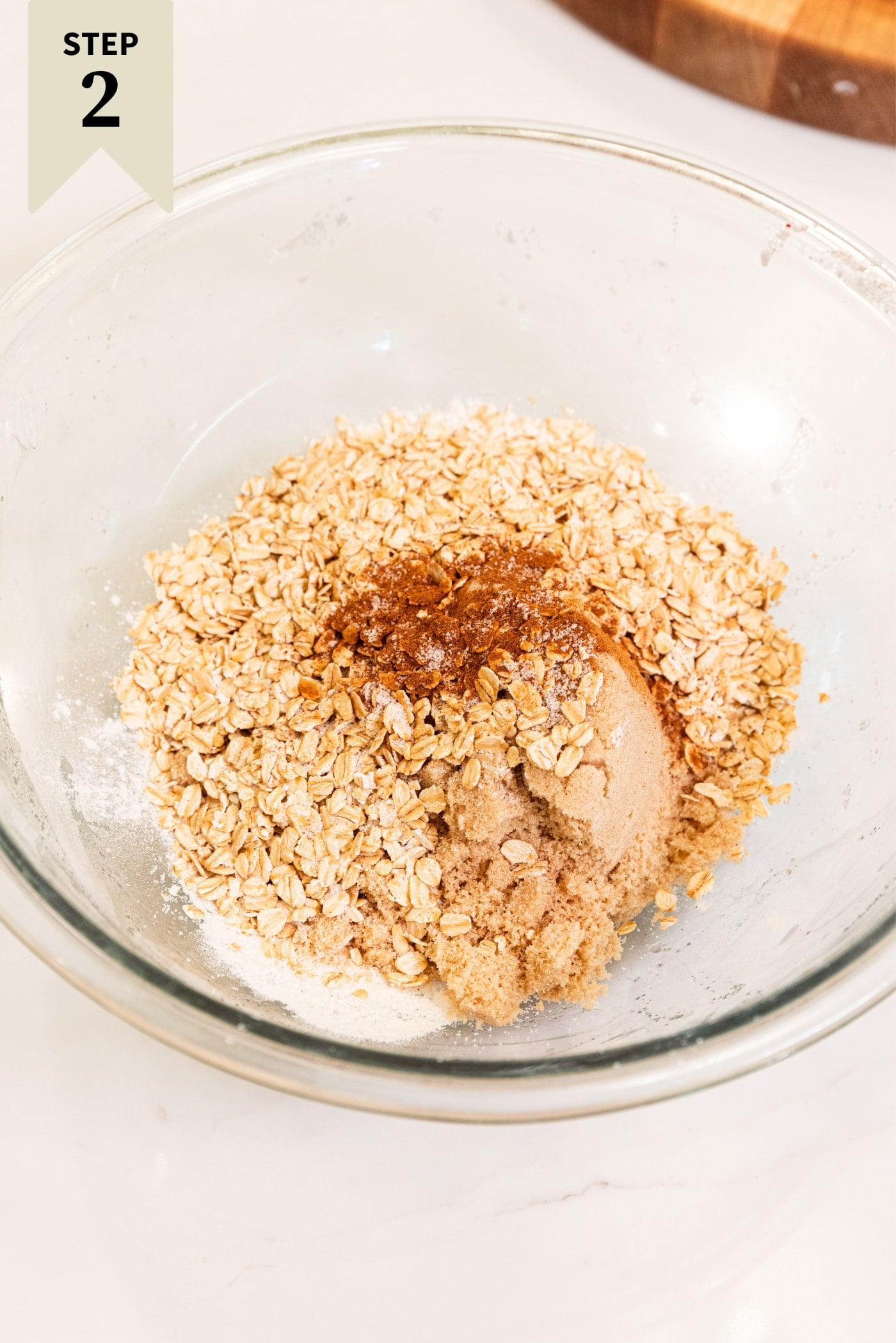 Glass bowl filled with combination of flour, cinnamon, brown sugar, and oats before being mixed together all on white surface.