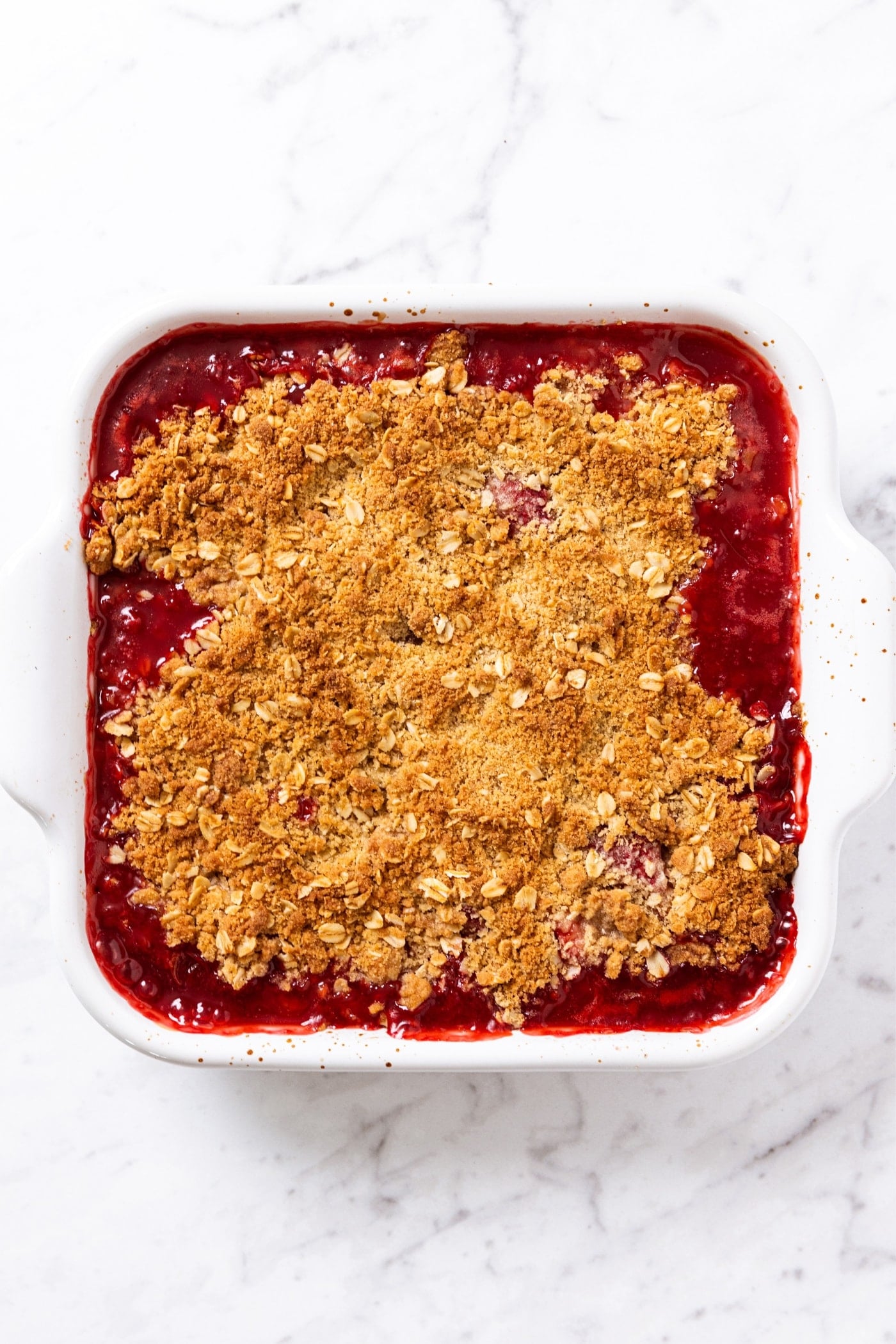 White baking dish sitting on white marble surface with baked strawberry rhubarb crumble right after coming out of the oven showing red bubbling mixture coming over the side of the crumble topping.
