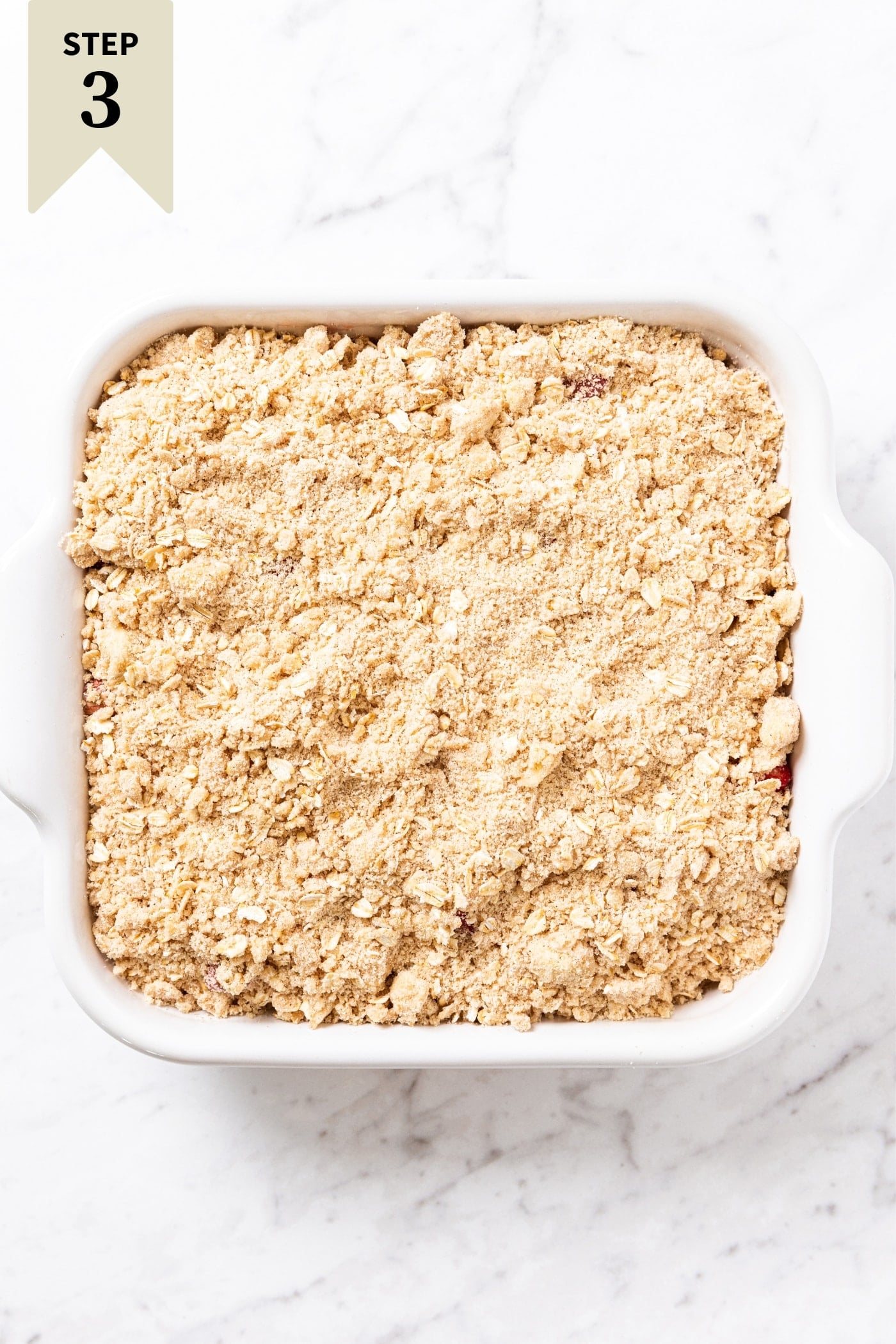 White baking dish sitting on marble surface filled with unbaked strawberry rhubarb crumble with light brown oat mixture on top.
