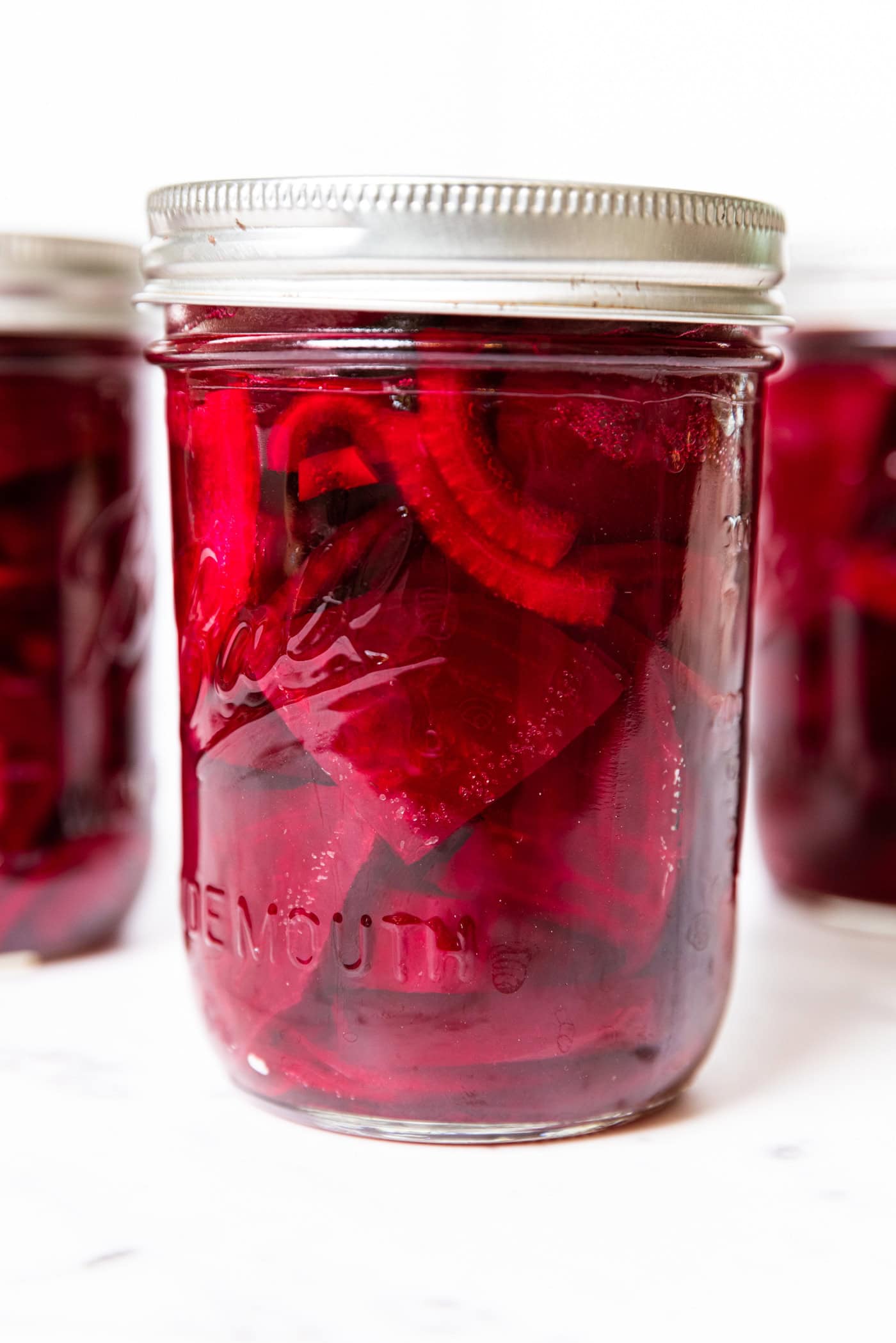 White surface filed three jars of pickled beets in red liquid with slices of onions.