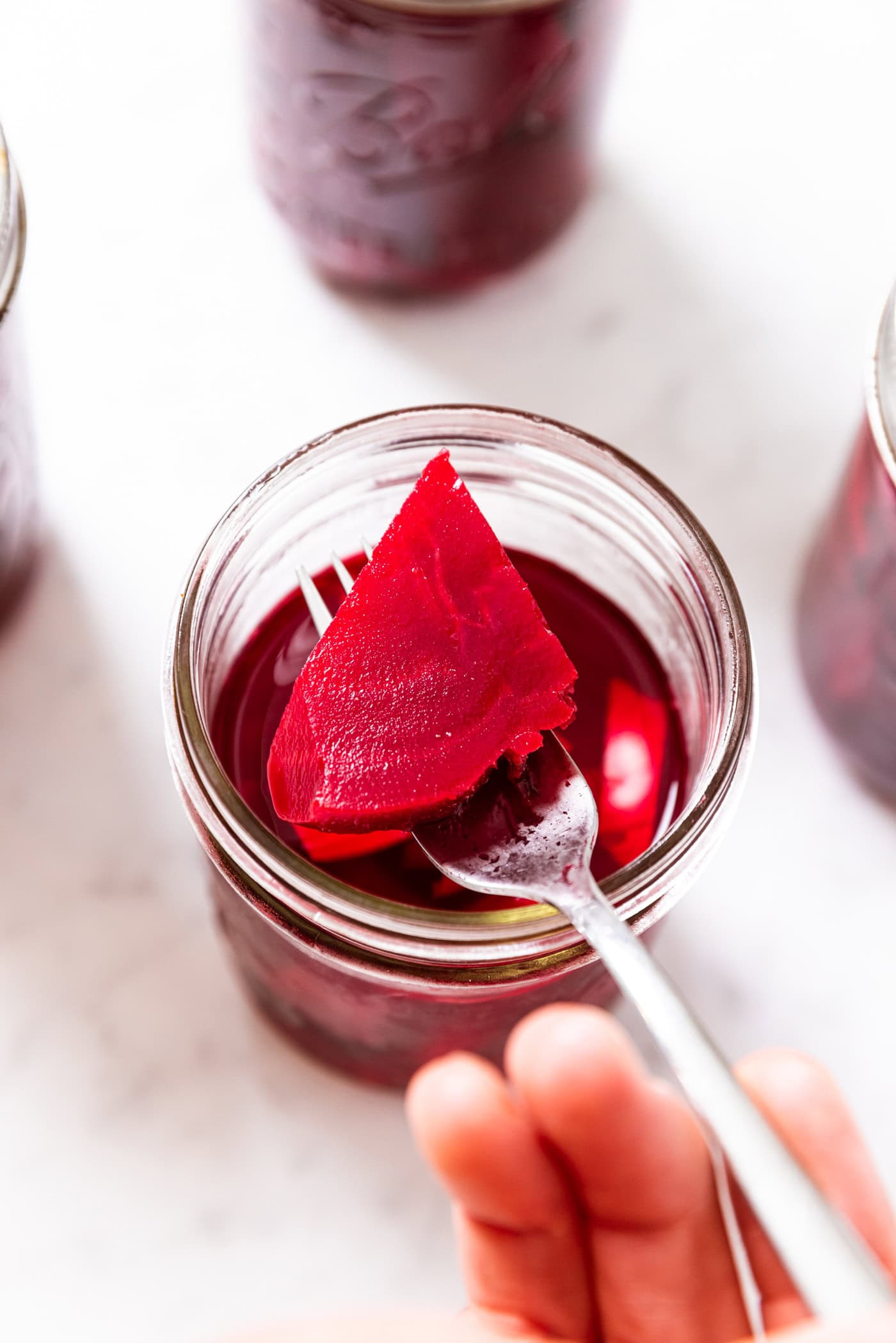 Hand holding fork with red colored slice of pickled beet above jar filled with liquid.