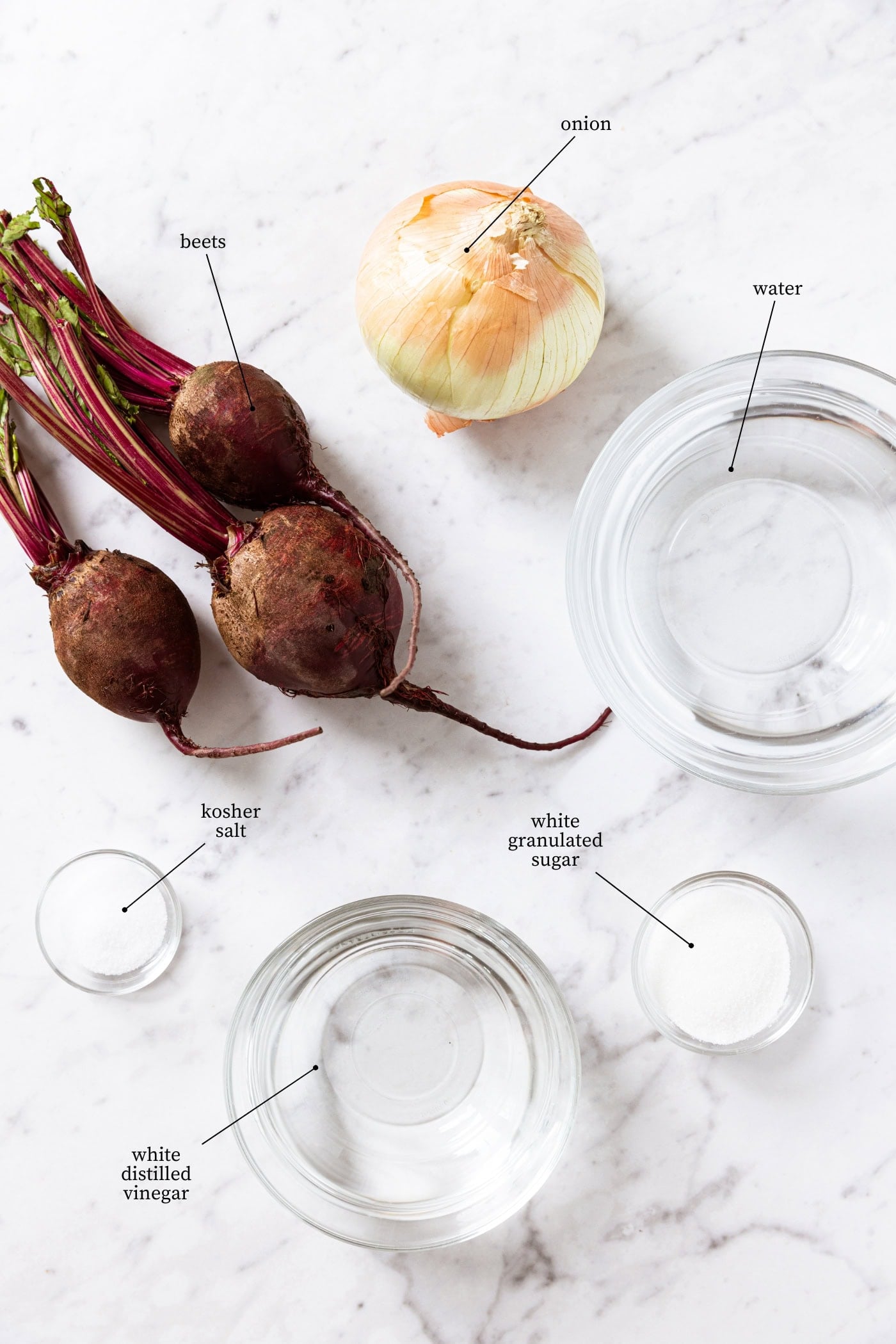 White marble surface with ingredients needed for pickled beets including raw beets, water, onion, sugar, salt, and vinegar.