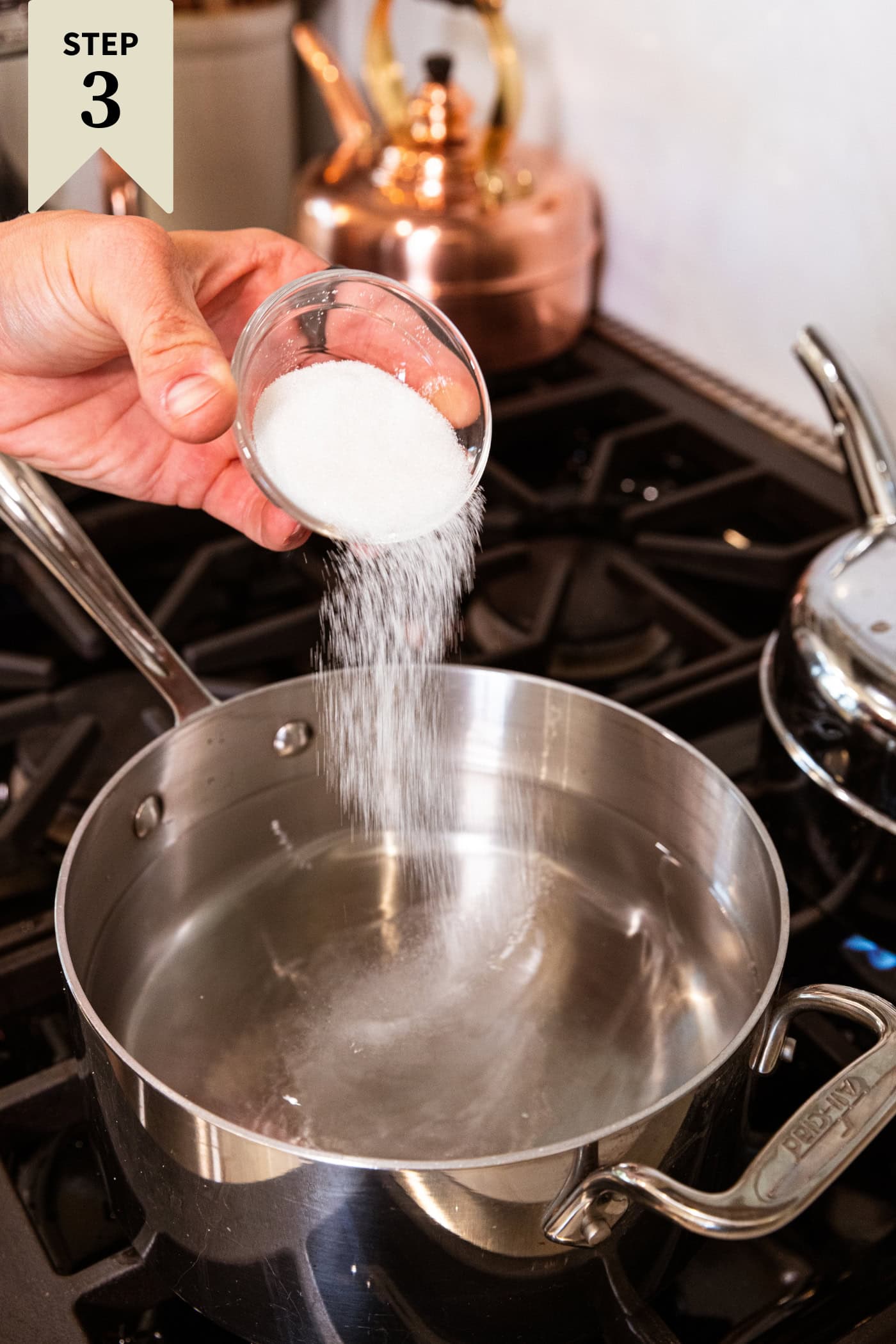 Hand pouring salt into a large saucepan filled with vinegar on the stovetop.