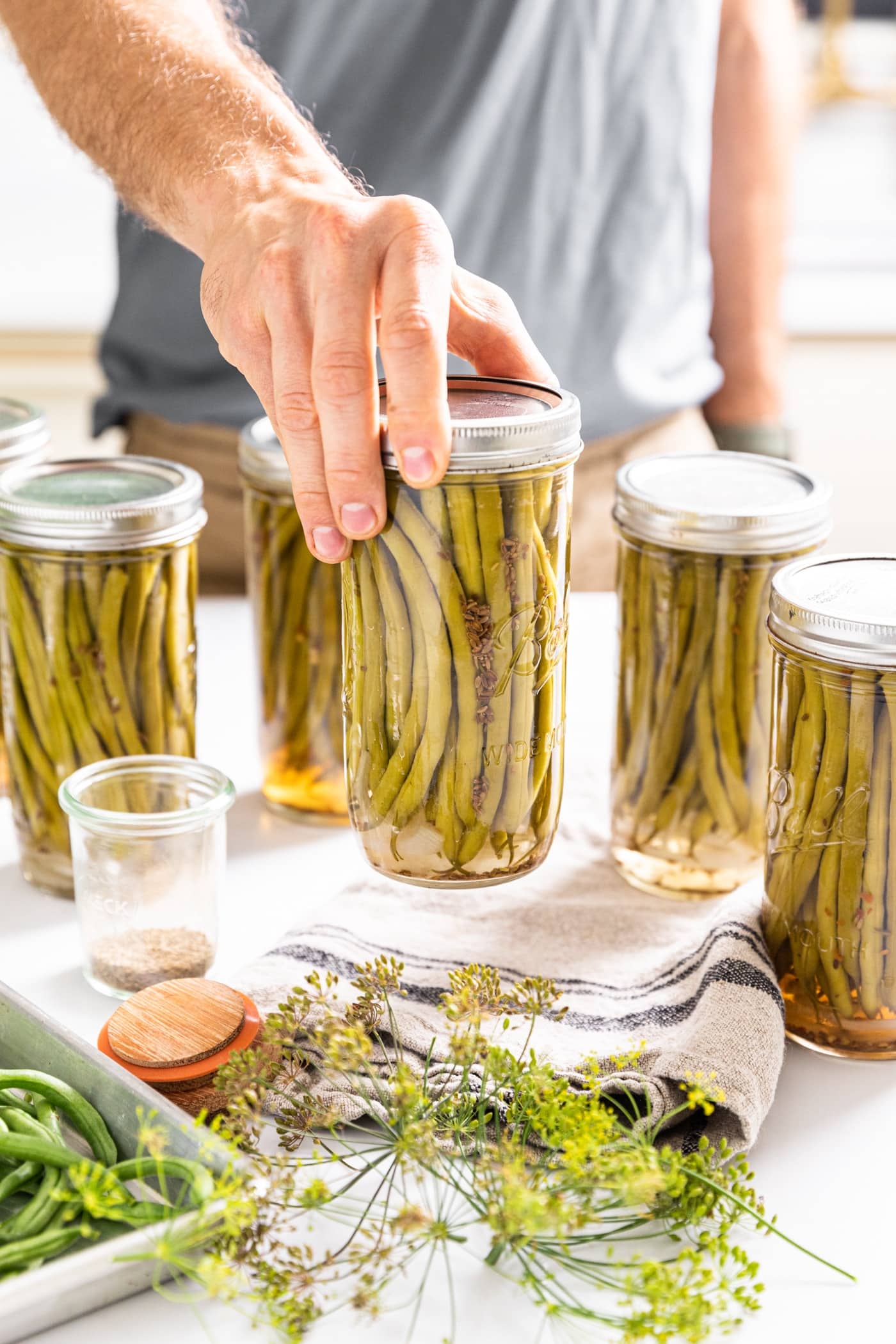 Hand holding glass jar filled with green beans with a towel underneath and extra filled glass jars around with raw green beans on pan