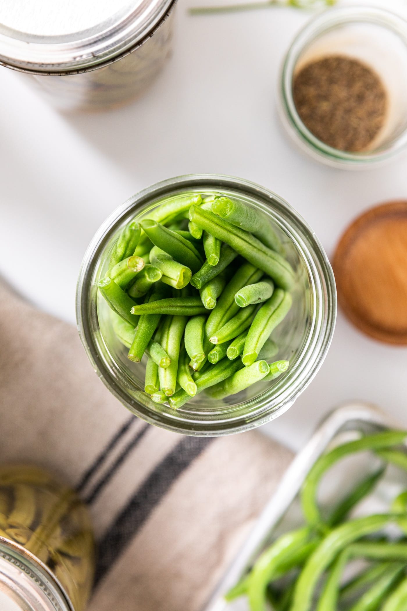 Top down view of glass jar filled with green beans with extra canning supplied all around on white countertop