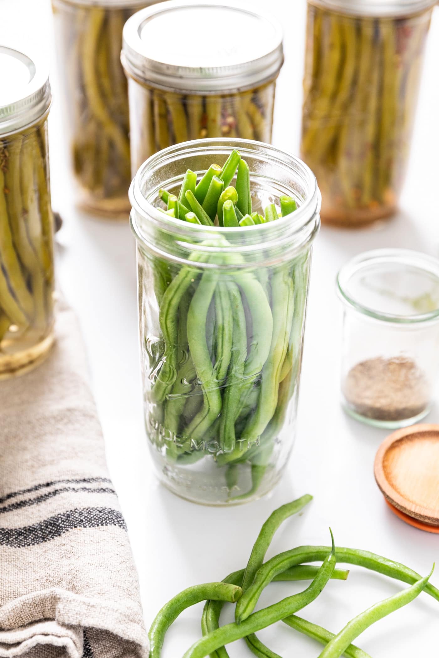 Glass jar filled with bright green beans without lid and ring on top sitting among processed glass jars all on white countertop