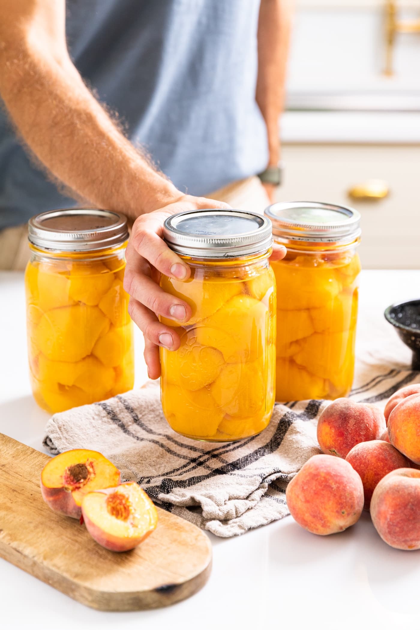 Hand holding large glass jar filled with slices of peaches laying on top of each other with other glass jars and unsliced peaches all around all on white countertop