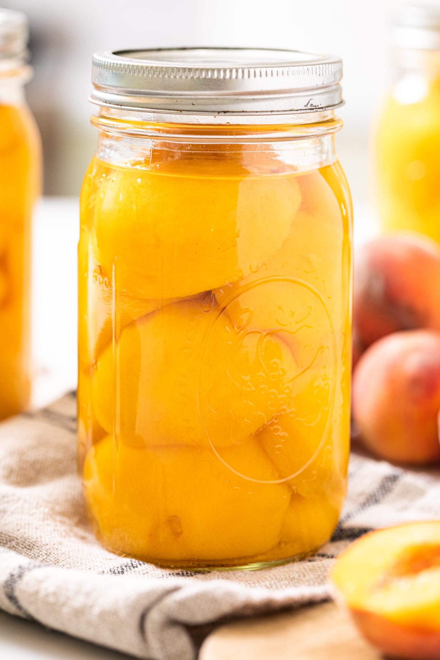 Large glass jar sitting on taupe colored cloth with slices of peaches sitting in clear liquid with peaches outside jar as well