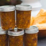 Five canning jars filled with peach chutney with bowl filled with bread in background all on gray slate surface