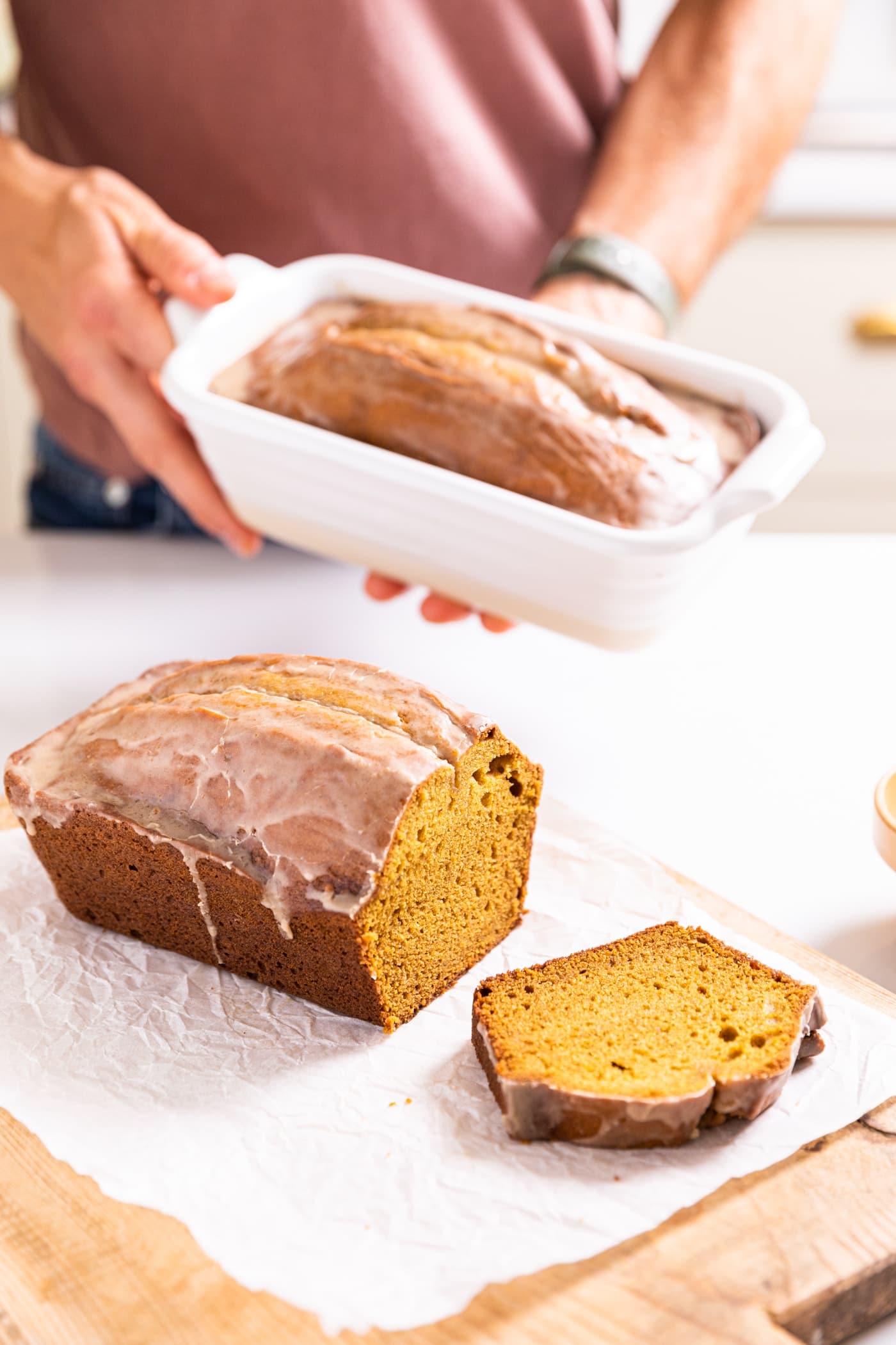 Hand holding pumpkin loaf in white loaf pan with sliced bread in front all on white countertop