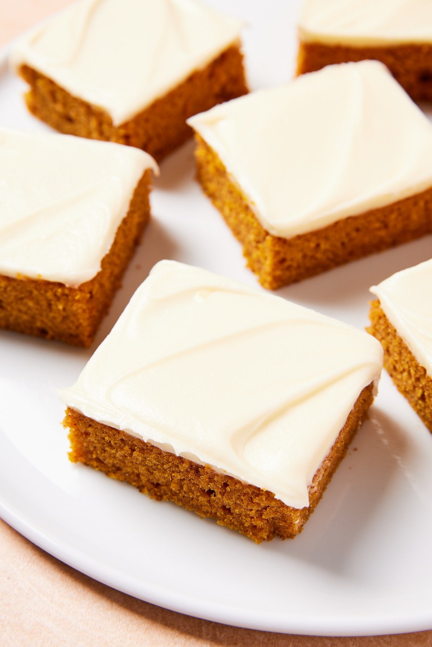 Squares of pumpkin bars with cream cheese frosting on top resting on round white plate.