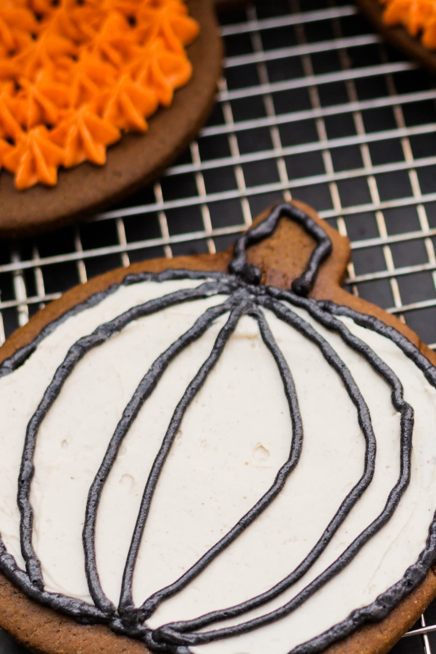 Spiced ginger cutout cookies topped with white frosting sitting on wire cooling rack with extra cookie with orange frosting in background