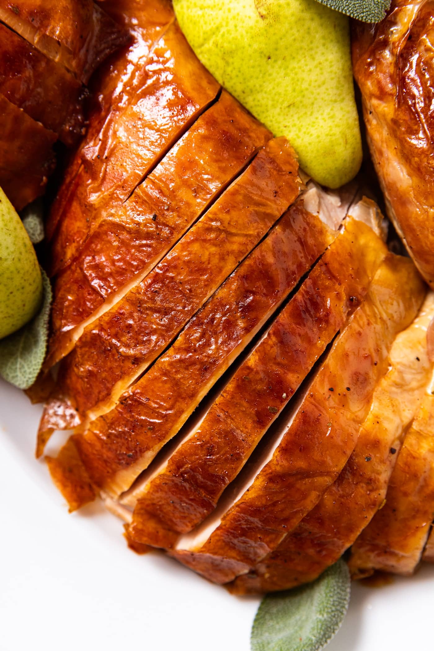 Slices of turkey breast sitting on white serving platter with pears and sage leaves all around.