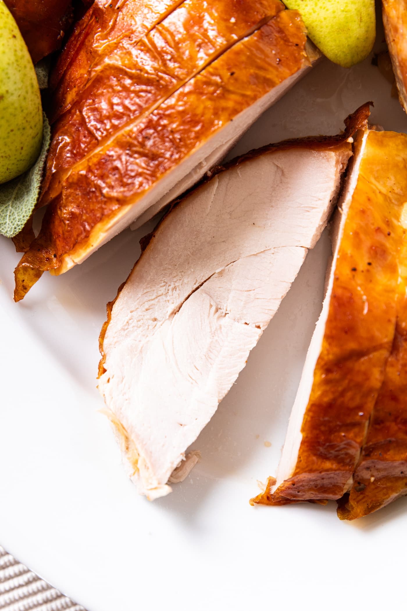 Slice of turkey breast sitting on its side amongst other slices of turkey breast on a white serving platter with pears and sage all around.