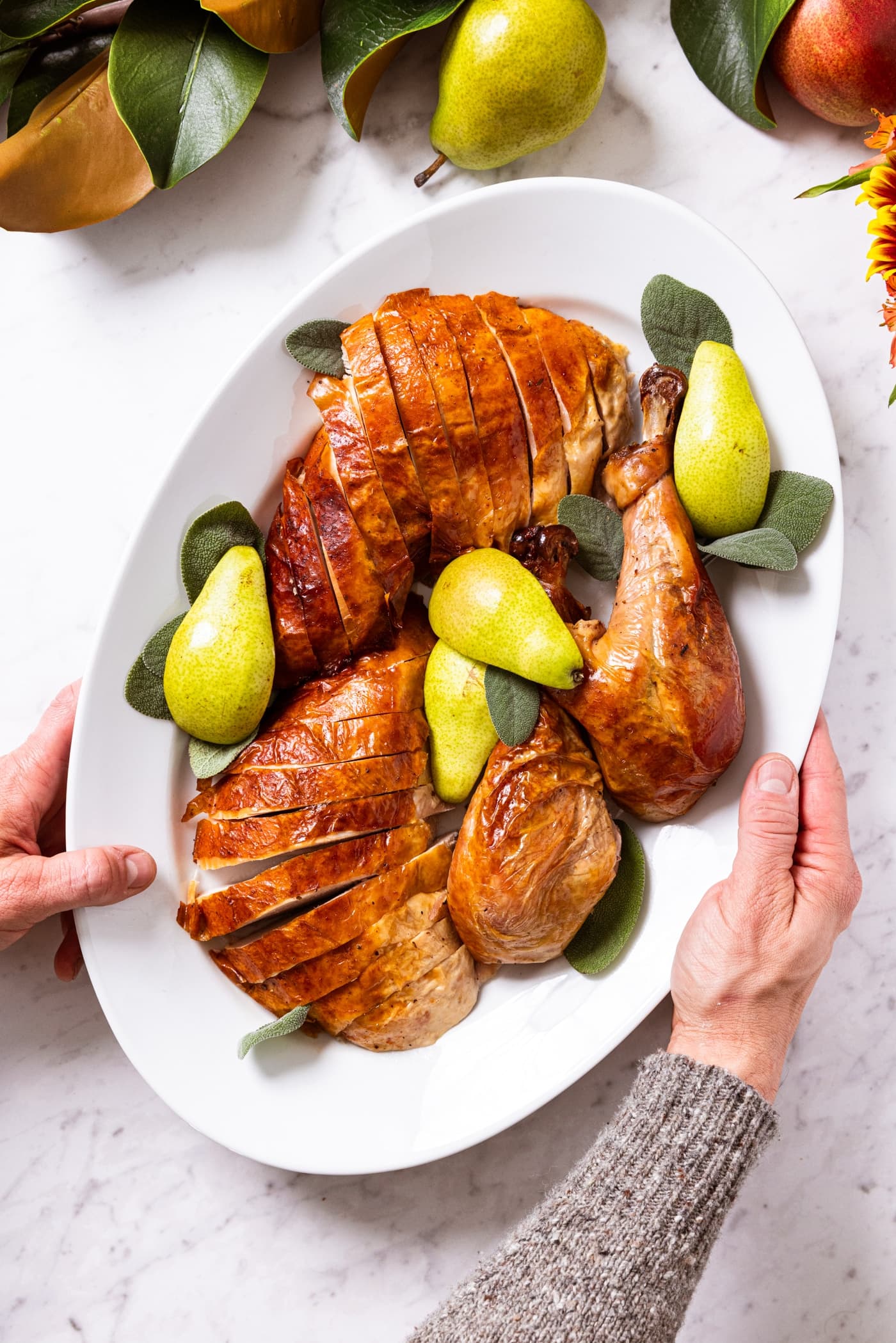 White serving platter filled with classic turkey that was sliced into parts including the breast, thighs, and legs with pears and sage leaves all around.
