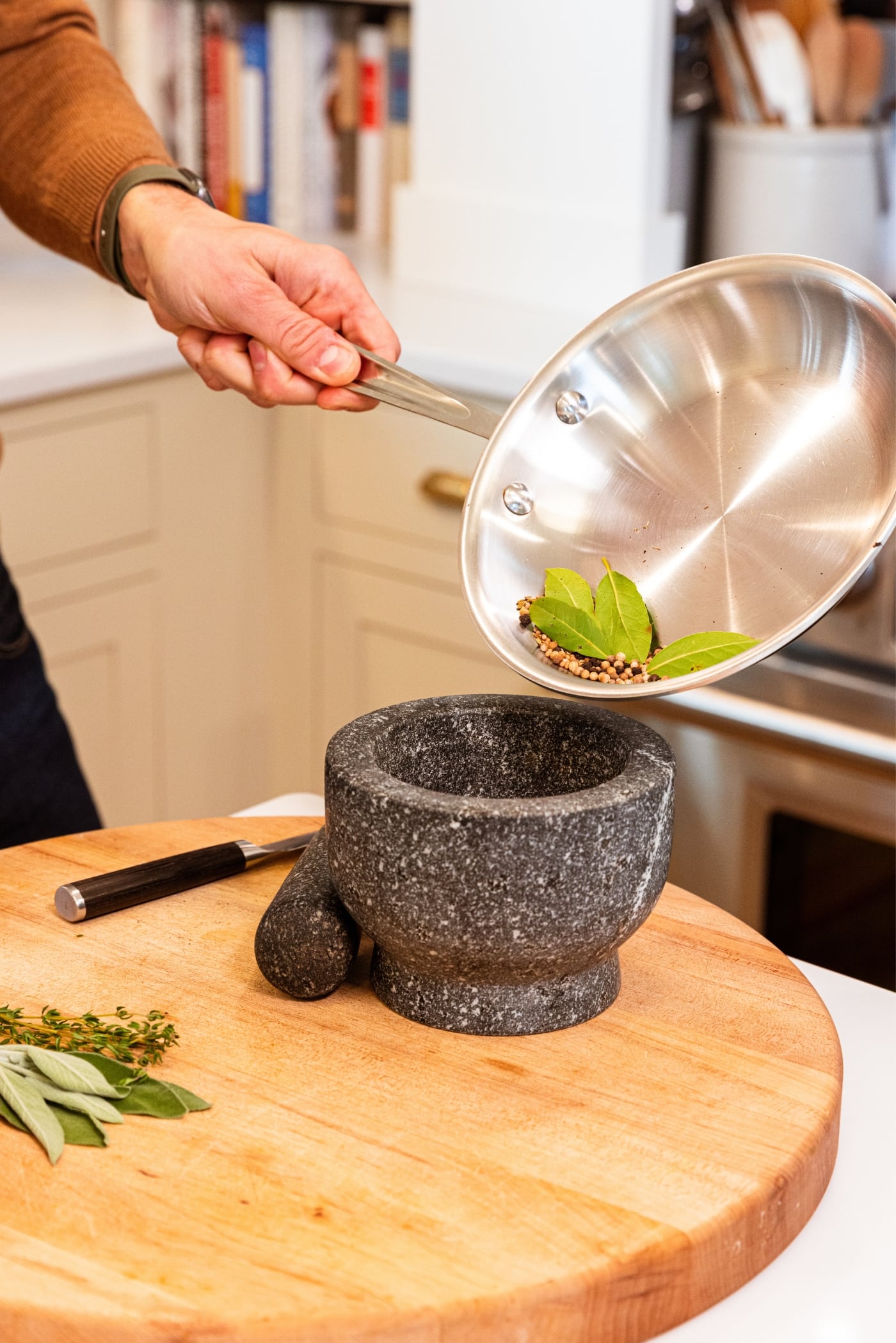 Hand holding metal skillet pouring contents into a black color mortar on wood cutting board with toasted spices in skillet.