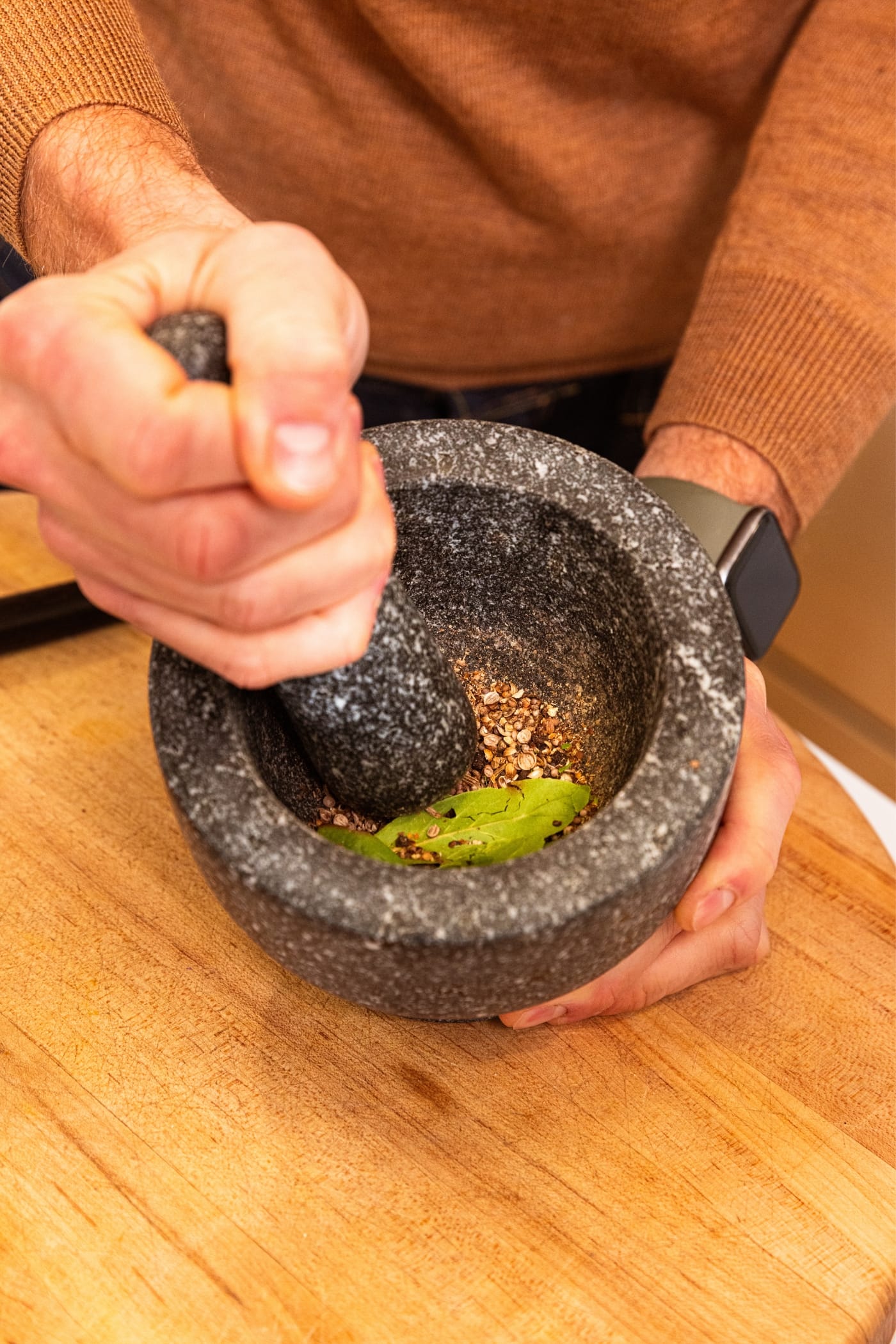 Hand holding pestle grinding contents of black colored mortar filled with spices and bay leaves with a wooden cutting board underneath.