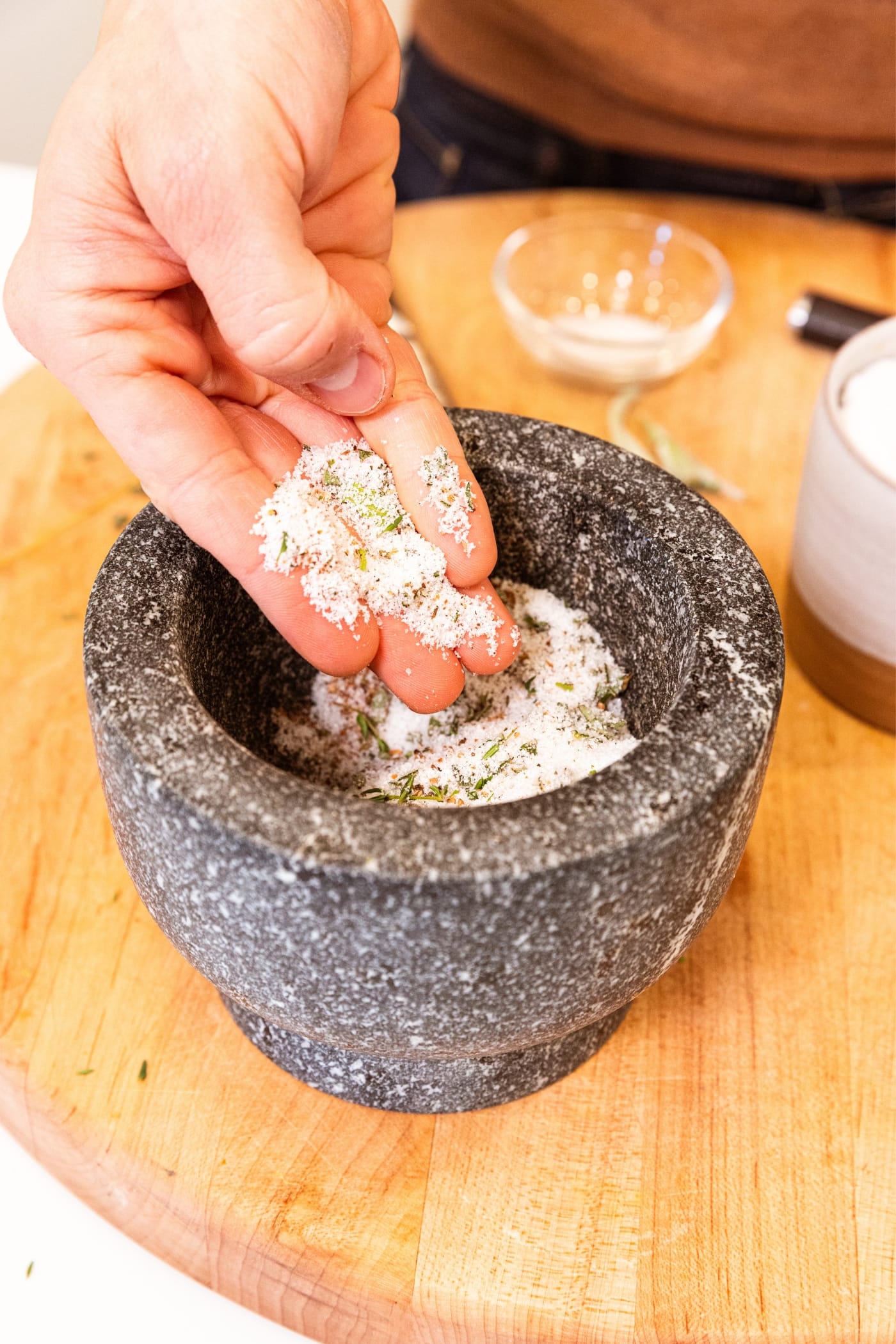 Hand holding sugar and salt mixture with crushed spices mixed in with contents in a gray colored mortar on wood cutting board.