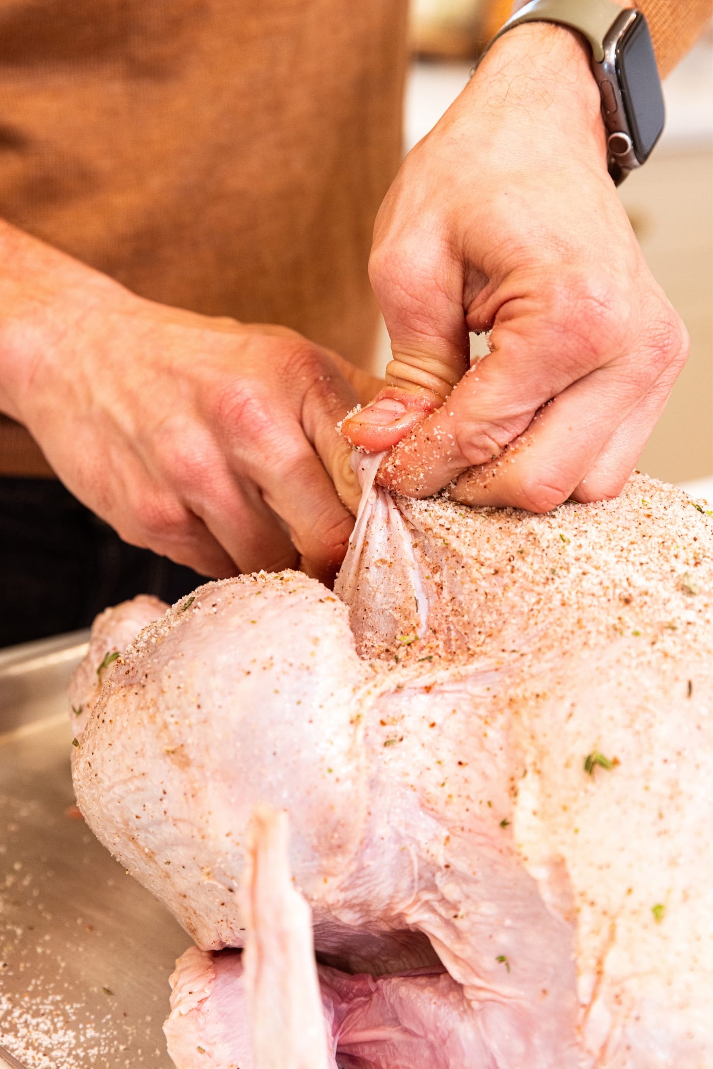 Hand pulling back skin from raw turkey with salt dry-brine mixture all over the top of the turkey.