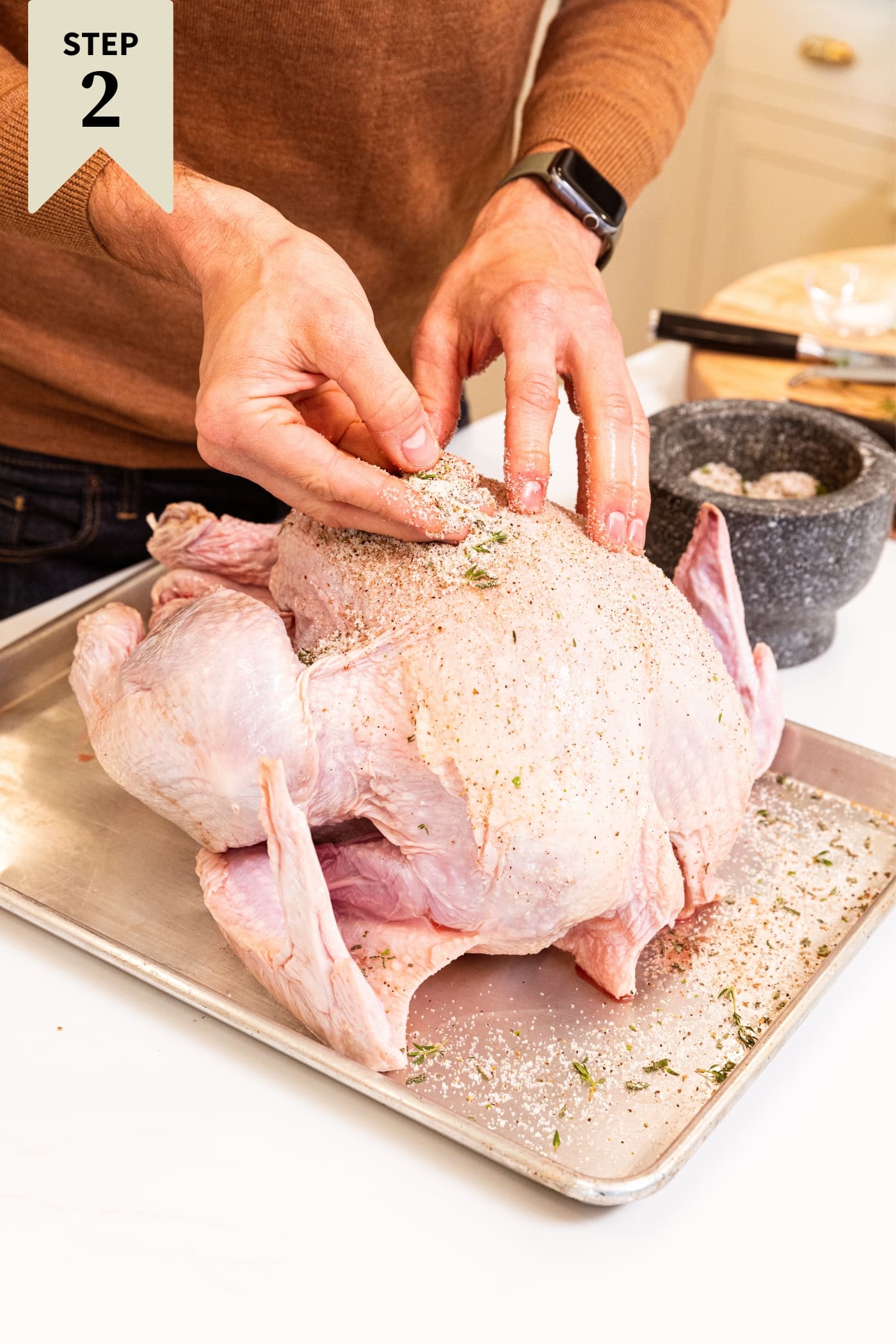 Hand placing salt mixture as a dry-brine all over the top of a raw turkey sitting on a metal baking sheet.