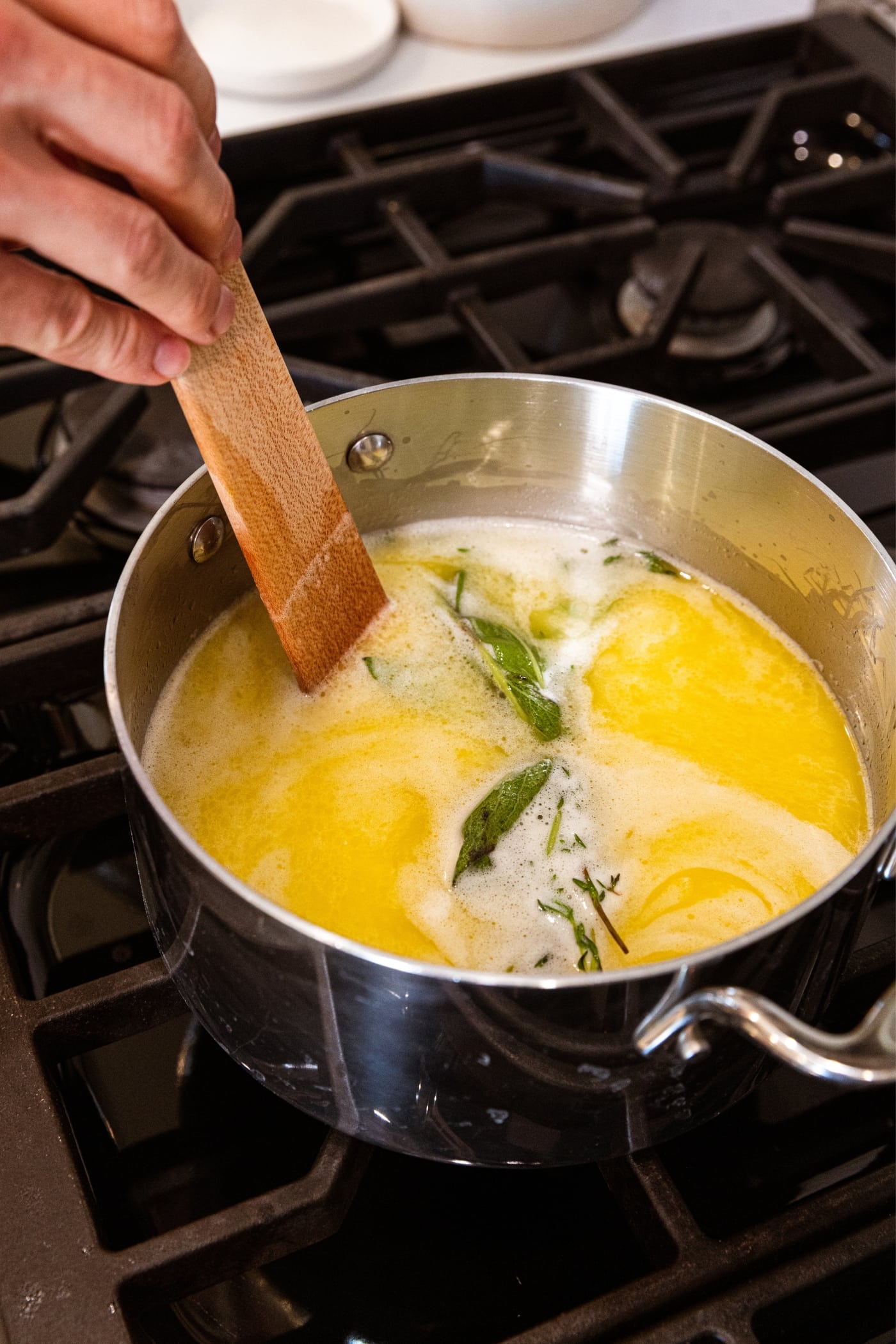 Wooden spatula stirring together herb packet tied together with twine sitting in butter and white wine mixture in metal kettle on stovetop.