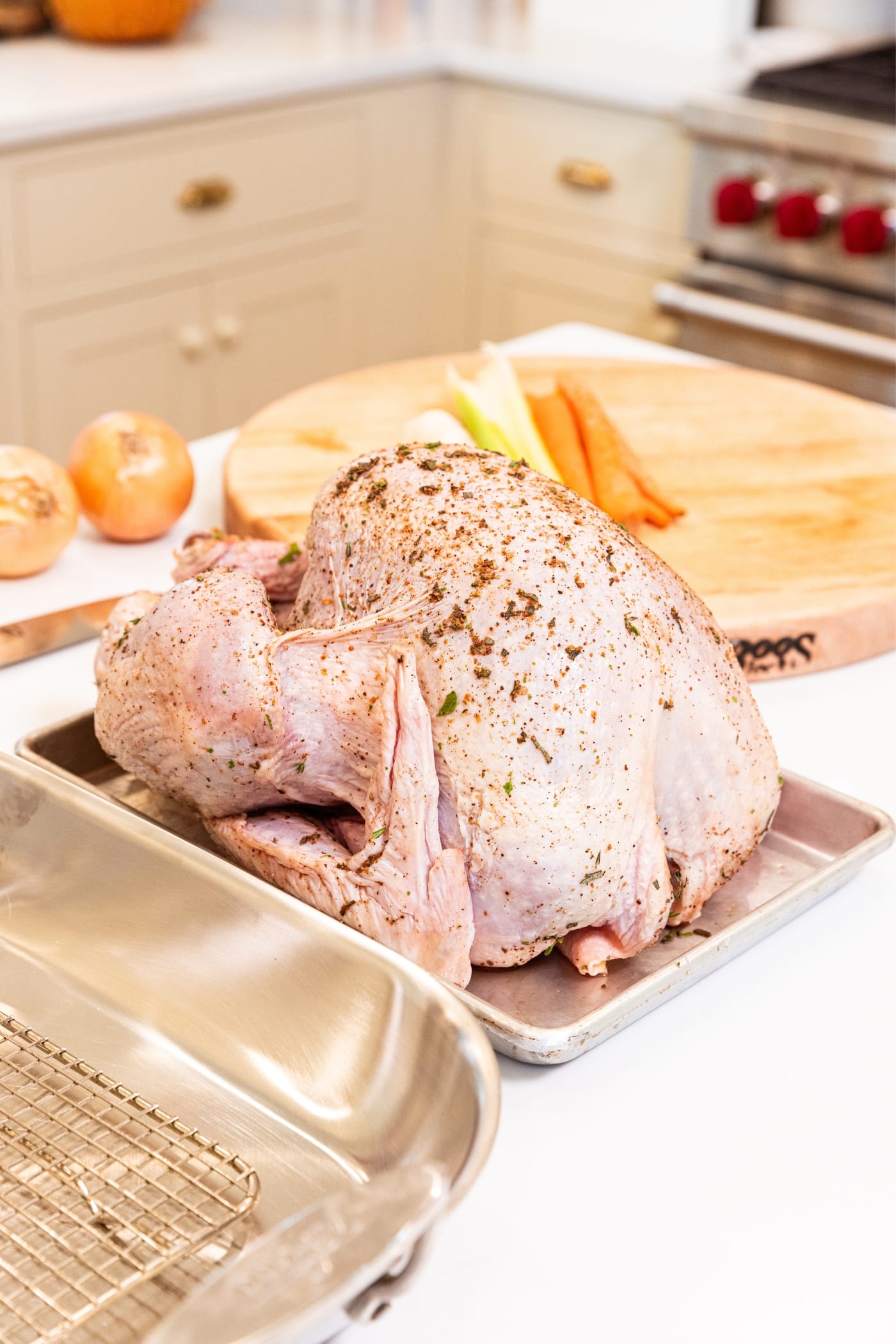 Raw turkey sitting on metal baking pan with herbs and dry-brine mixture all over the top with carrots and celery and onion on wood cutting board in the background.