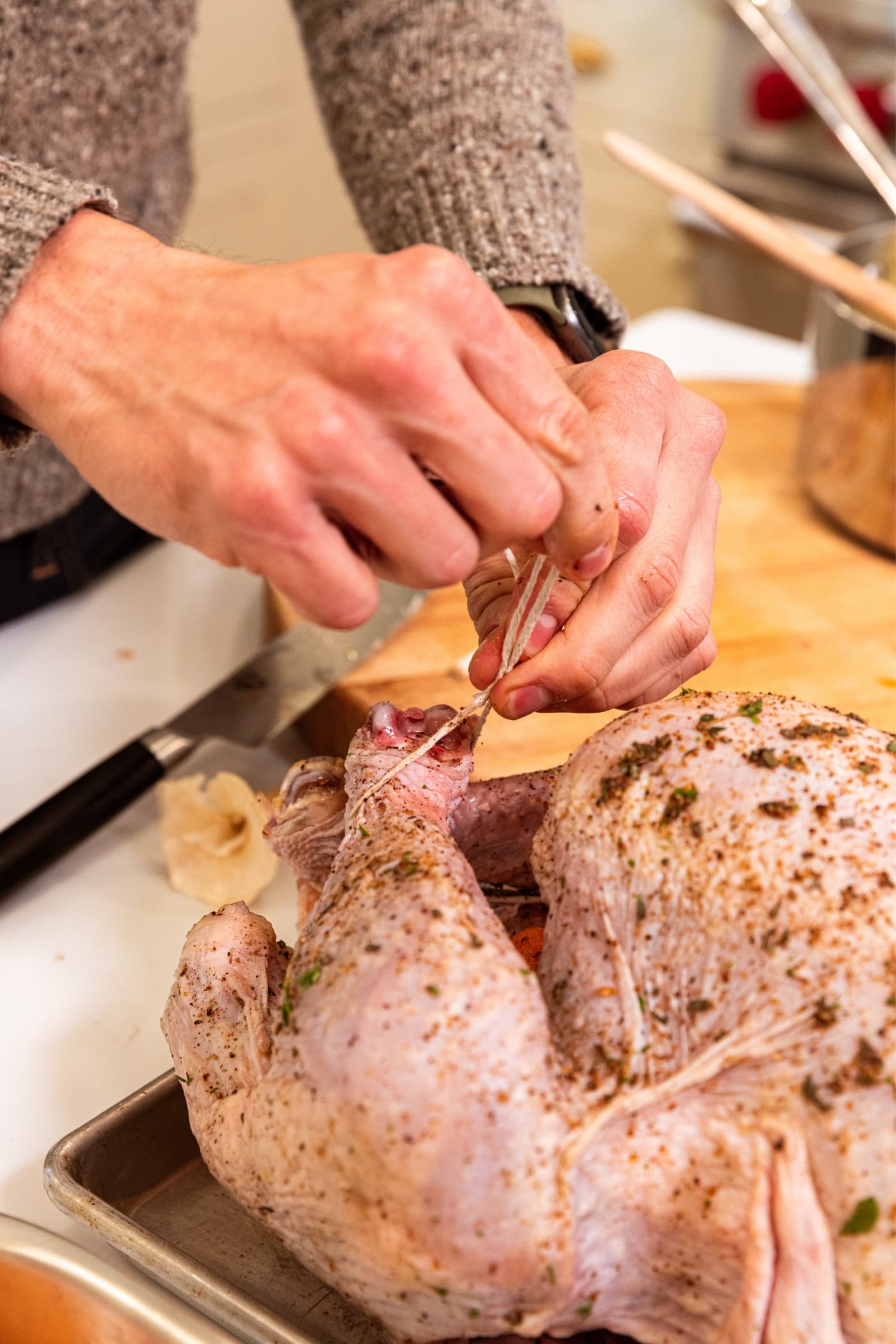 Two hands trussing up turkey legs with butcher's twine with turkey covered in a dry-brine on metal rack.