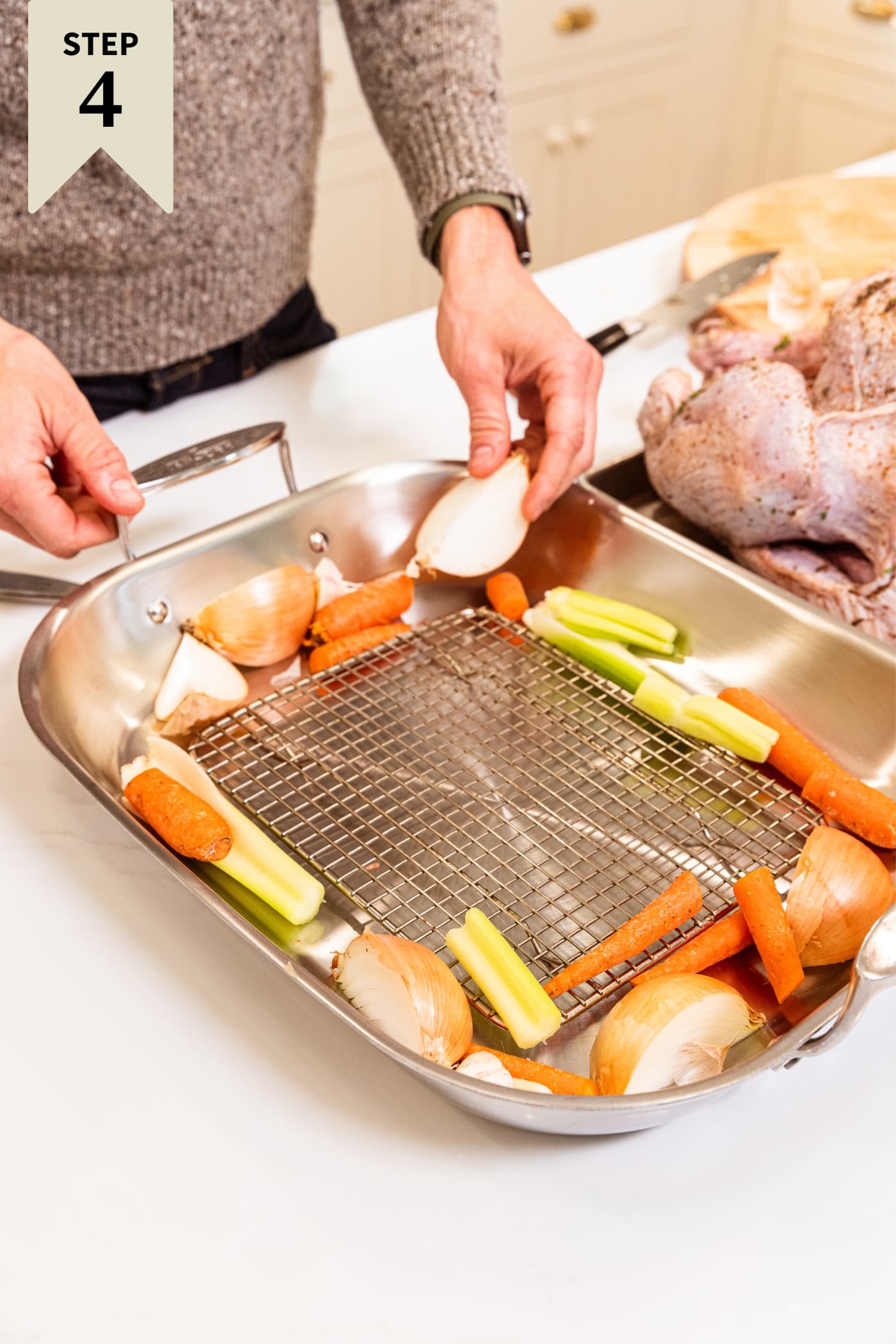 Hands placing onion and celery and carrots in a metal roasting pan fitted with a metal rack in the bottom with raw turkey to the side before being placed on rack.