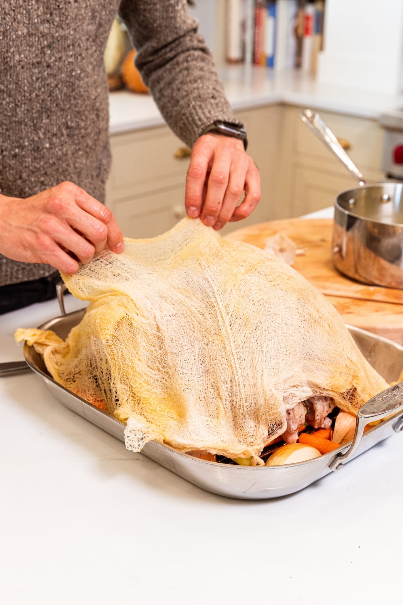 Hands placing saturated cheesecloth on top of raw turkey sitting on metal rack in roasting pan with carrots and celery and onion resting all around.