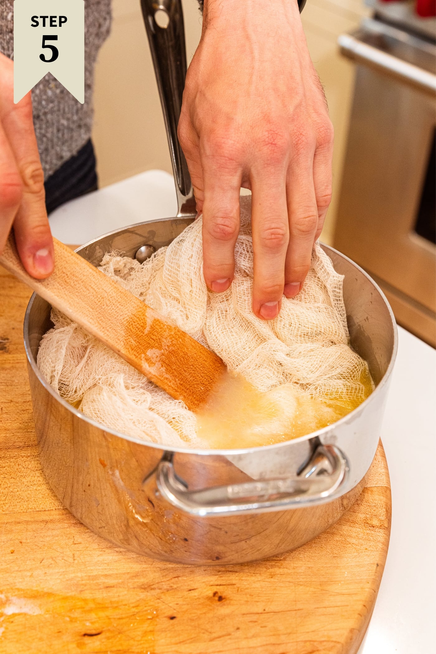 Hand and wooden spatula pushing cheesecloth into yellow colored basting liquid in metal kettle sitting on wooden cutting board.
