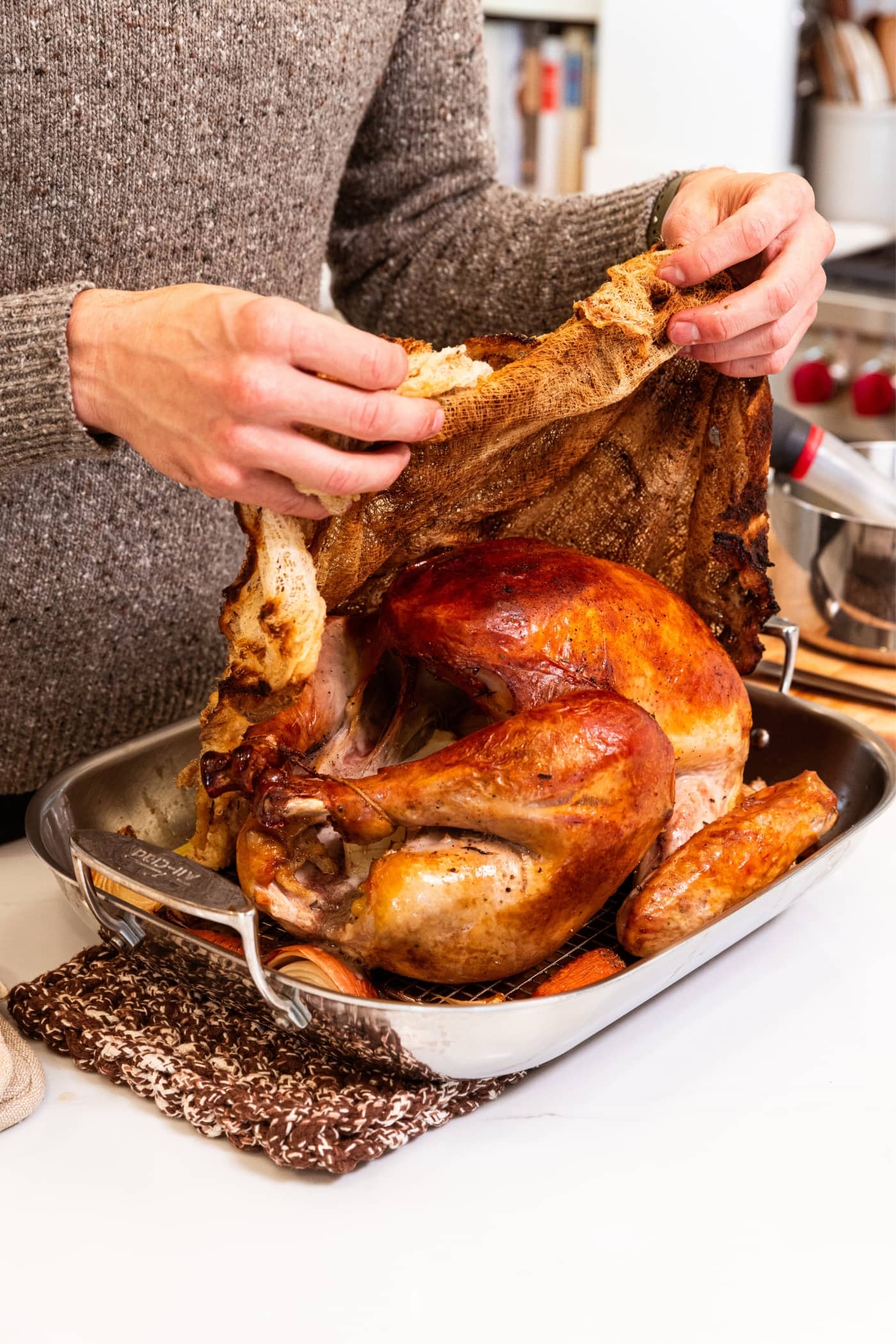 Hand removing cheesecloth from top of baked turkey breast sitting in metal roasting pan after baking in the oven.
