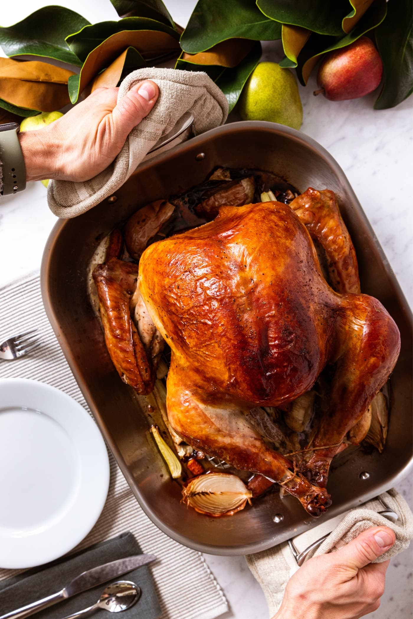 Two hands holding handles of roasting pan filled with baked turkey breast that's golden brown on top with plate sitting on placemat and magnolia garland.