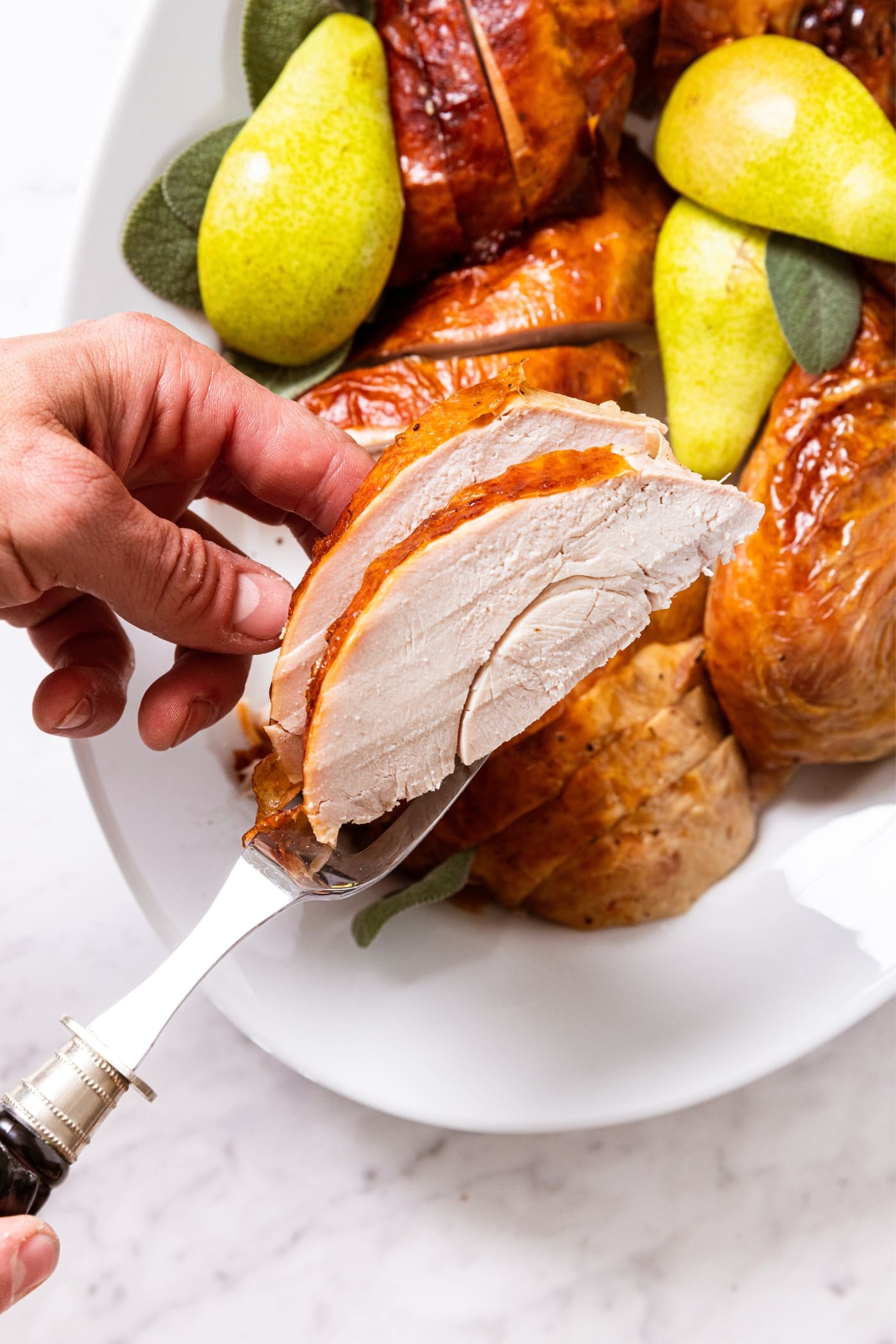 Hand holding two slices of turkey breast over a serving platter filled with rest of turkey with serving prongs holding turkey.
