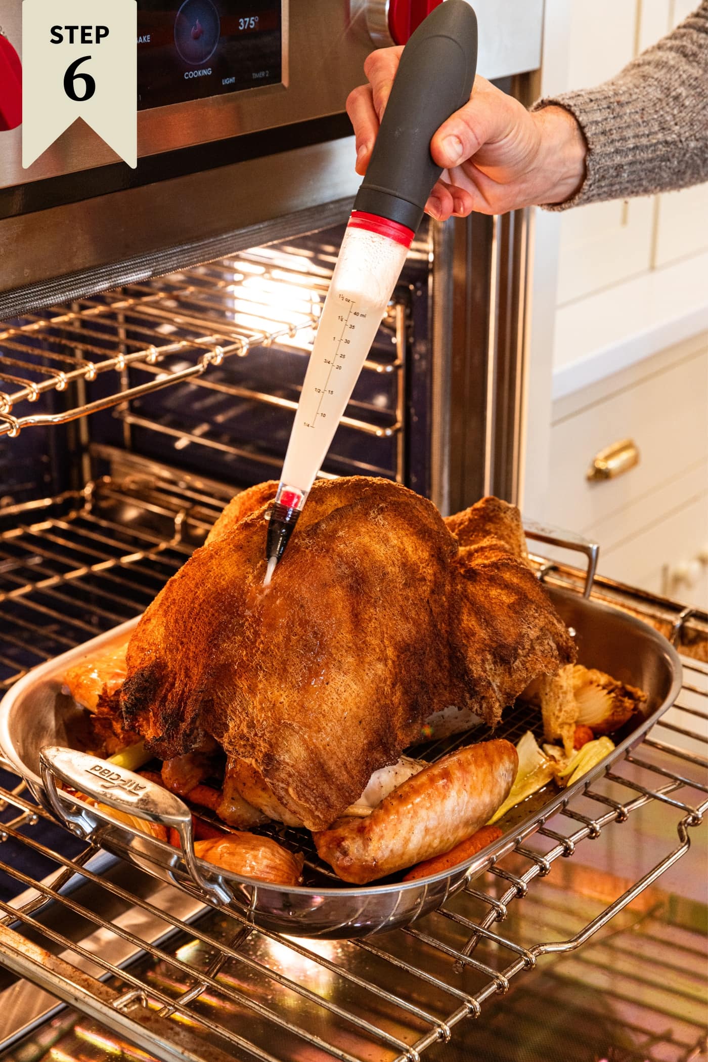 Hand using a baster to baste a baking turkey sitting on a rack that was pulled out of the oven with the oven door open.