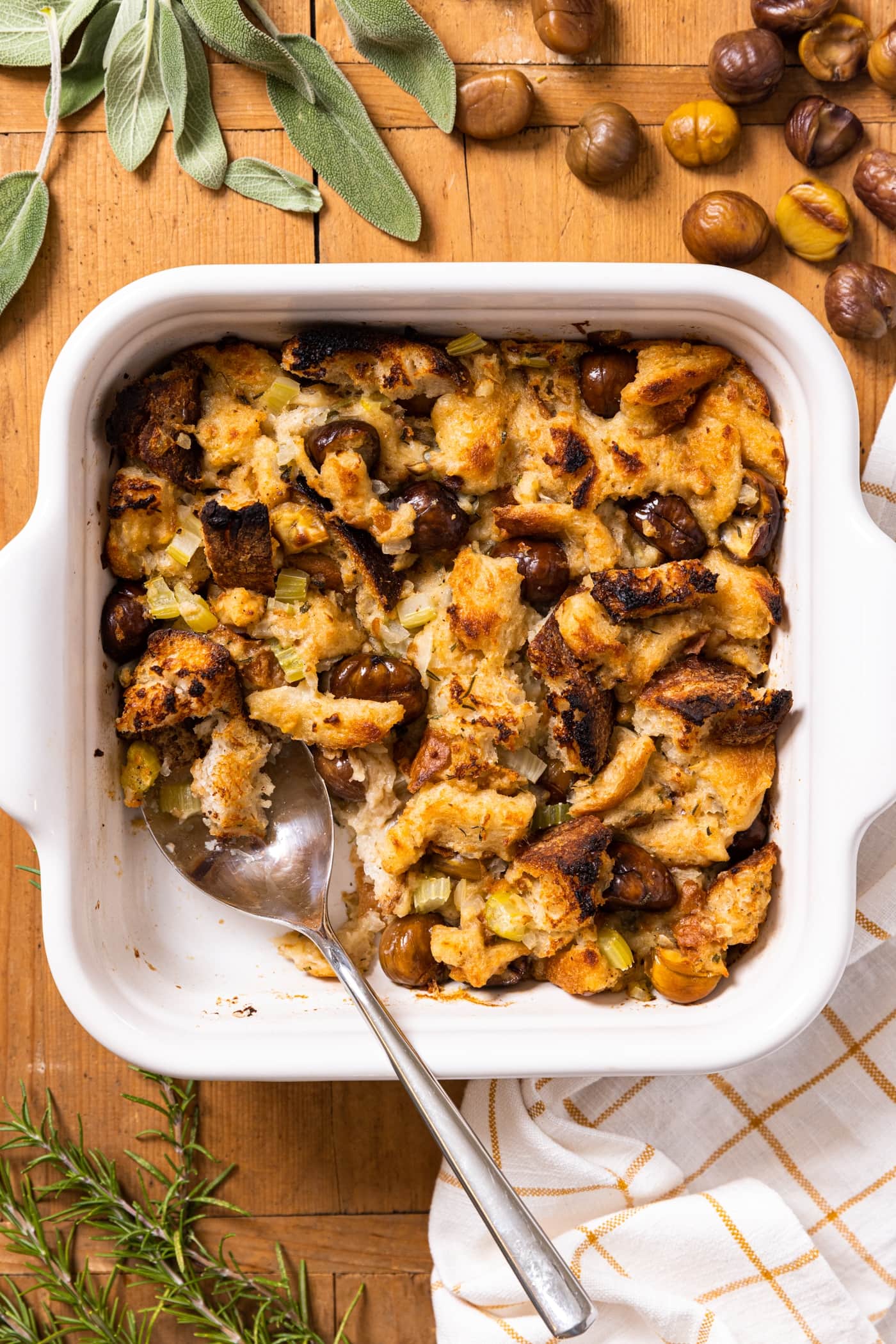 Top down view of white baking dish filled with slow cooker stuffing with spoon sitting inside with herbs and chestnuts all around on wood surface