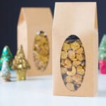Brown bags filled with caramel nut crispix sitting on white surface with second bag in background with small Christmas trees around.