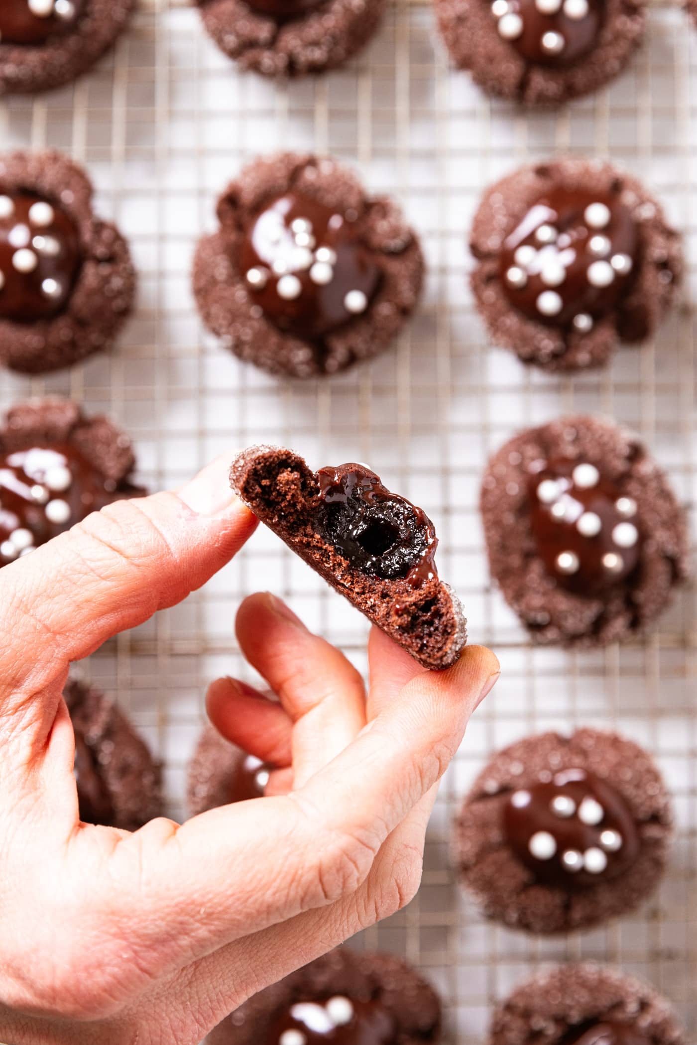 Hand holding chocolate cookie with bite taken out showing cherry inside of cookie with rest of cookies sitting below on a wire cooling rack.