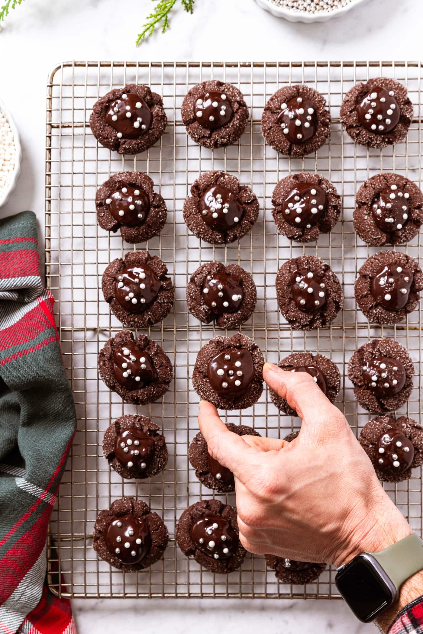 Hand placing one chocolate-covered cherry cookie on wire cooling rack filled with rest of chocolate cookies on white marble surface with red and green napkin to the side.