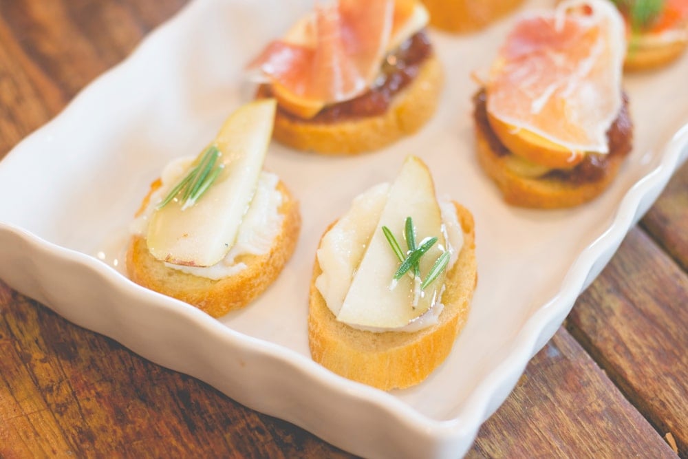 Crostini topped with ricotta and pear and sprig of rosemary sitting on white serving platter