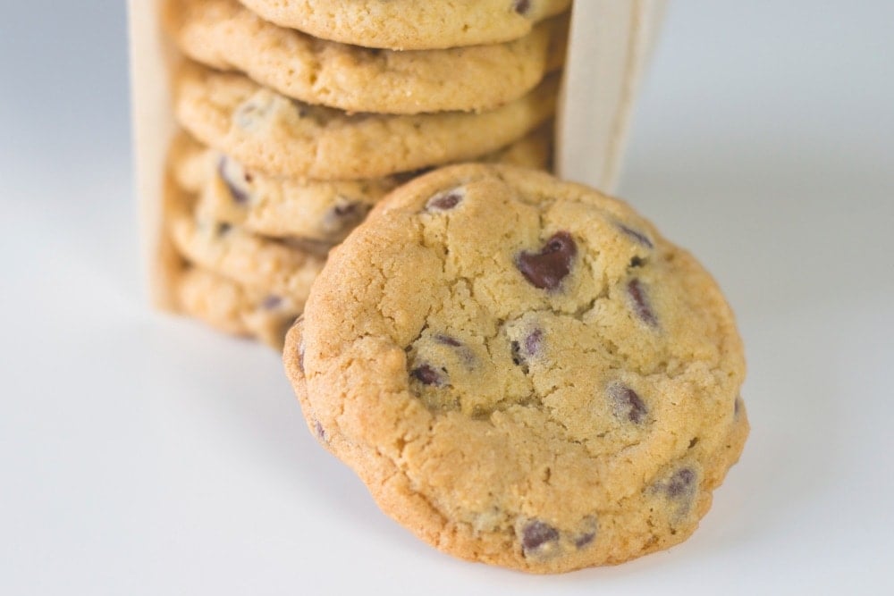 Stack of large chocolate chip cookies sitting on white countertop with one cookie in front and rest of cookies wrapped up in parchment in background