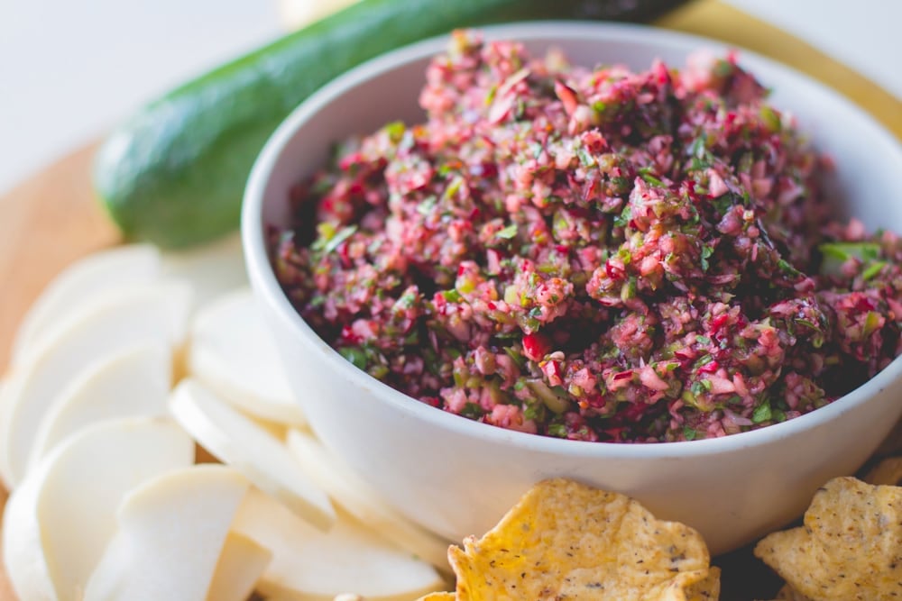 White bowl filled with red colored cranberry salsa with chips and sliced vegetables all around