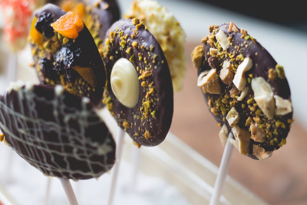 Several chocolate pops standing upright with different types of toppings including chopped nuts, dried fruit, and sprinkles