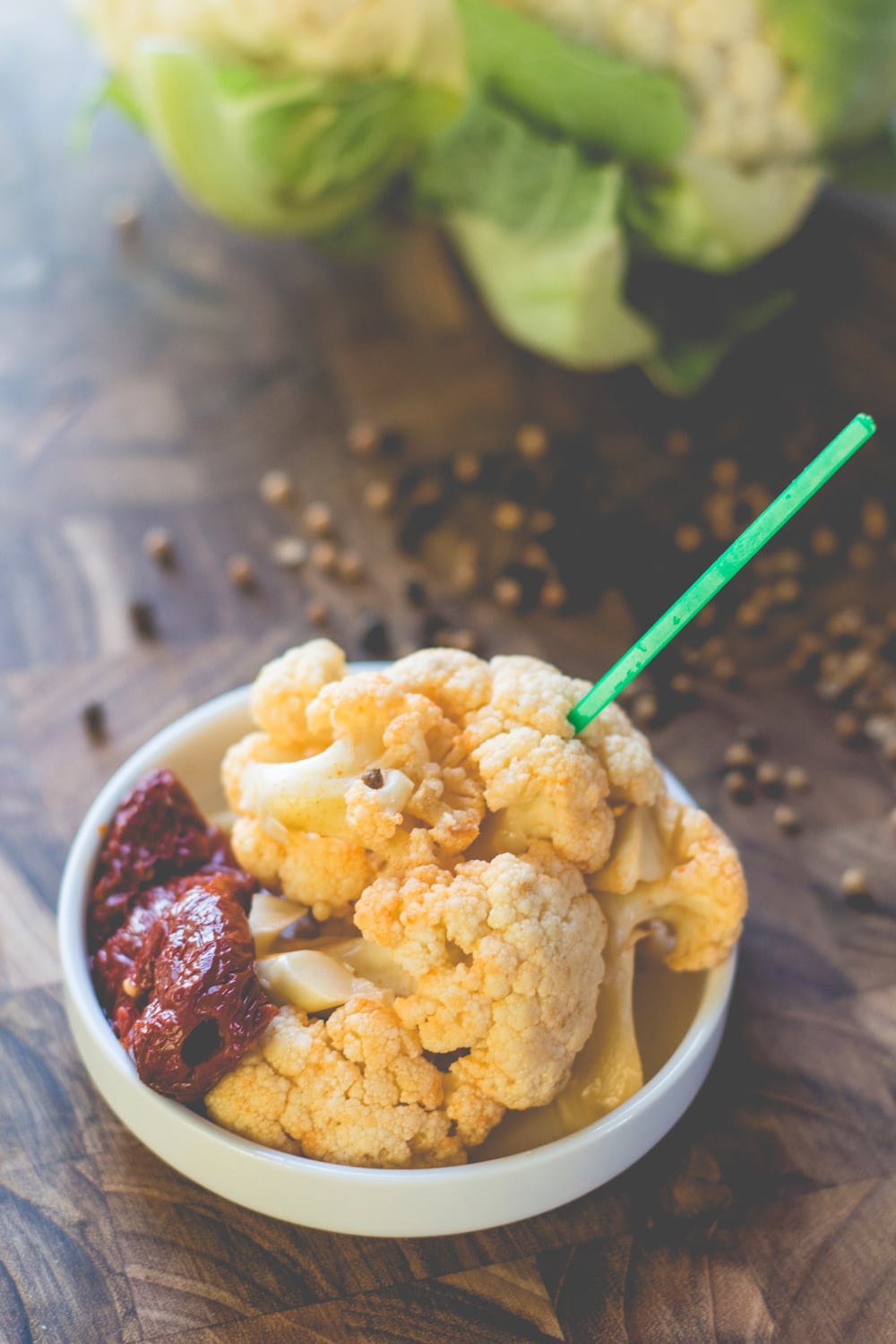 Small round plate filled with chipotle pickled cauliflower with red pepper sitting beside with green spear coming out of cauliflower with more cauliflower in background all on wood surface