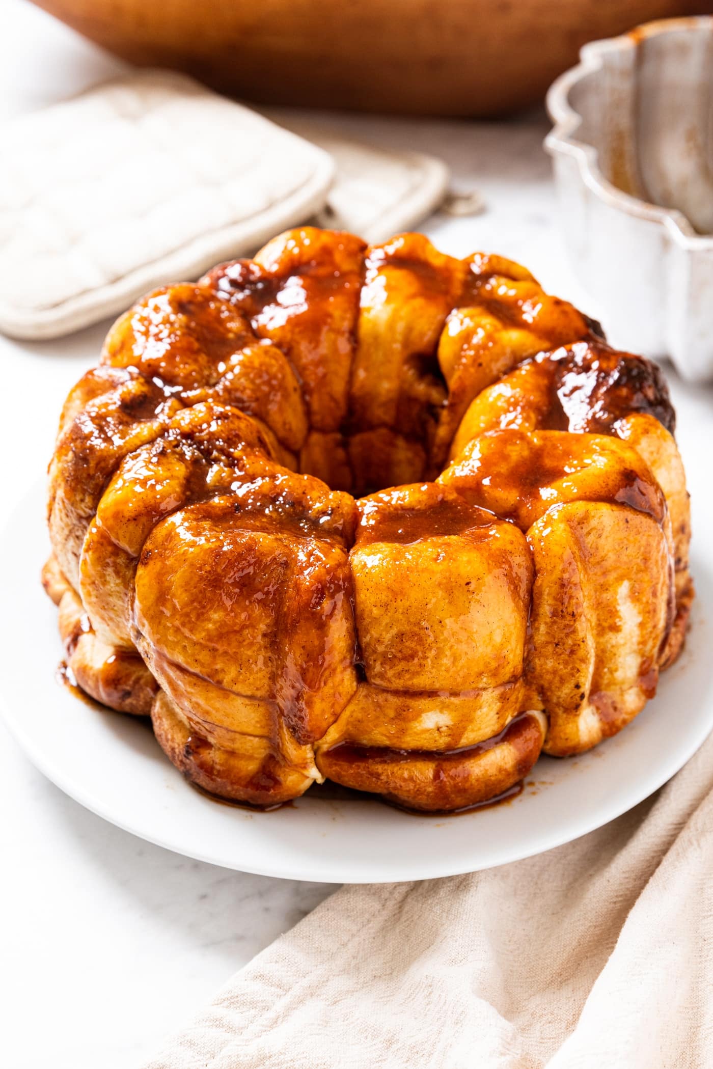 White plate filled with pieces of monkey bread with golden sauce all over the top with glistening edges all on white marble surface with Bundt pan in background.