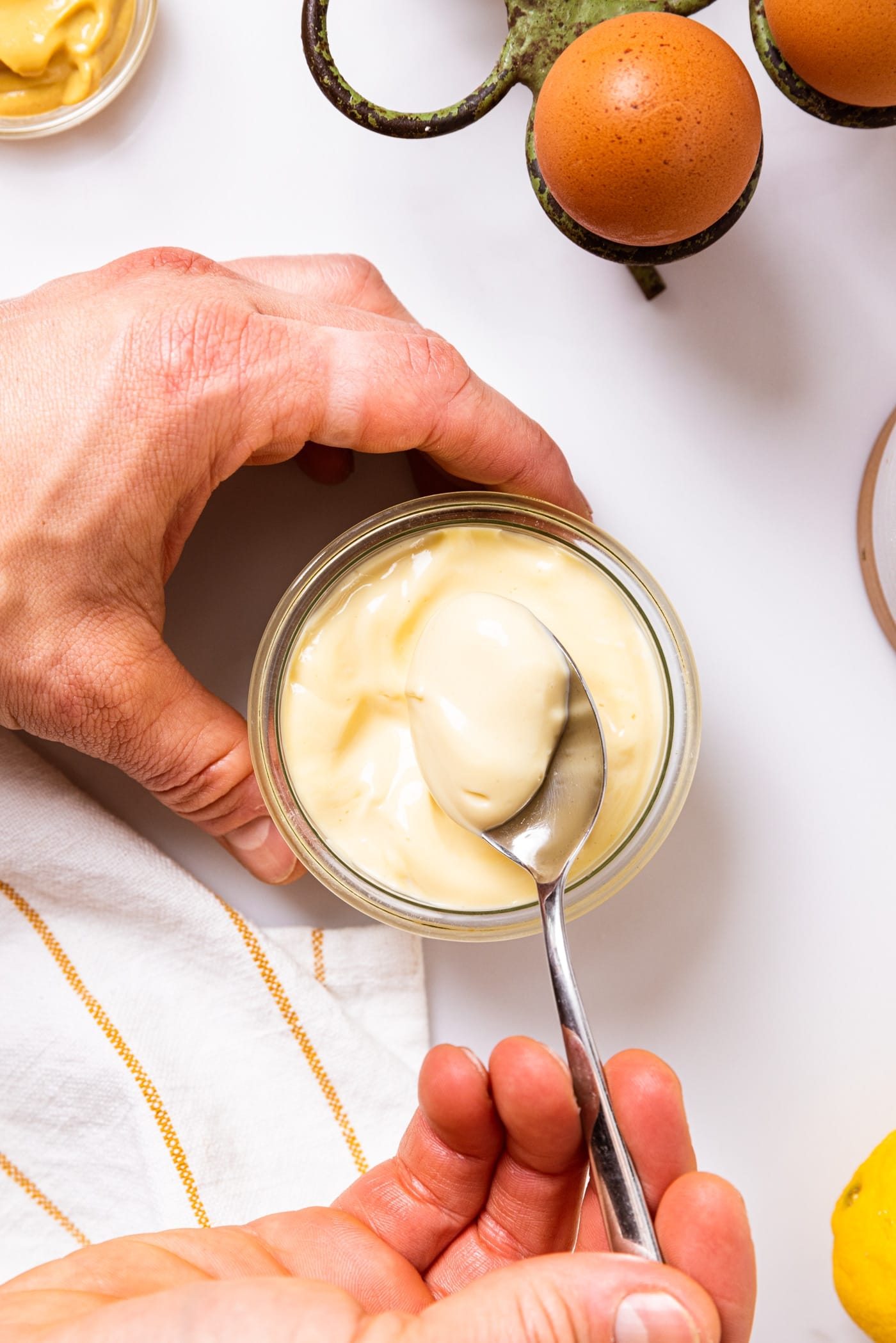 Top down view of two hands holding spoon scooping out homemade mayonnaise from glass bowl with lemons, eggs, and mustard on white countertop