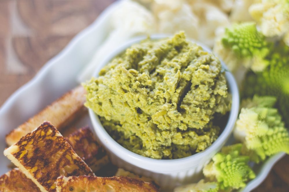 White bowl filled with chunky green-colored pea hummus with sliced vegetables and toasted bread sitting on wood board