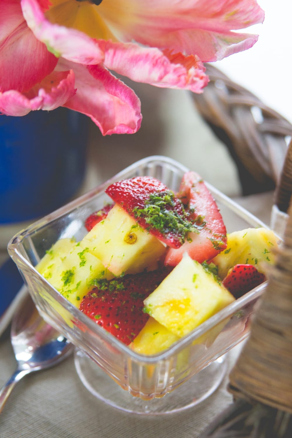 Small square bowl filled with fresh fruit including strawberries and pineapple with lime zest on top with flower in background