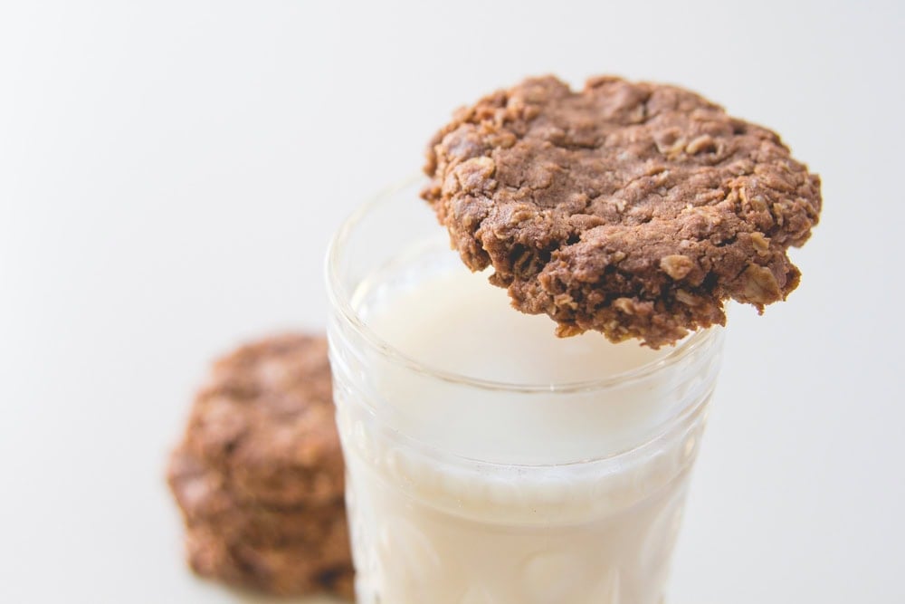 A double chocolate oatmeal cookie resting on the rim of a large glass of milk with more cookies to the side.