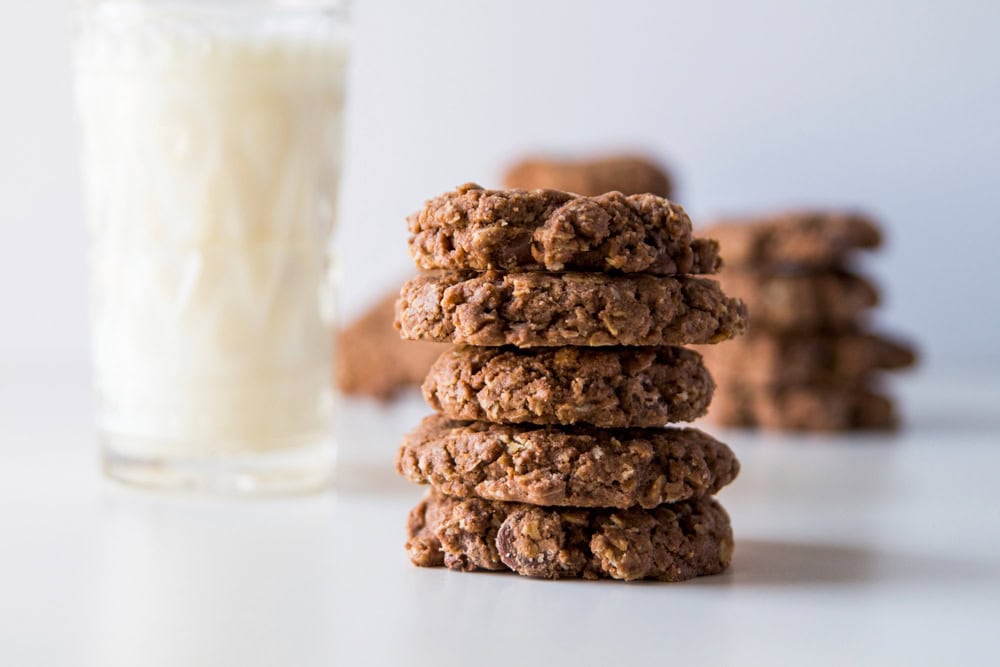 White surface with stack of double chocolate oatmeal cookies with more in the background along with a large glass of milk.