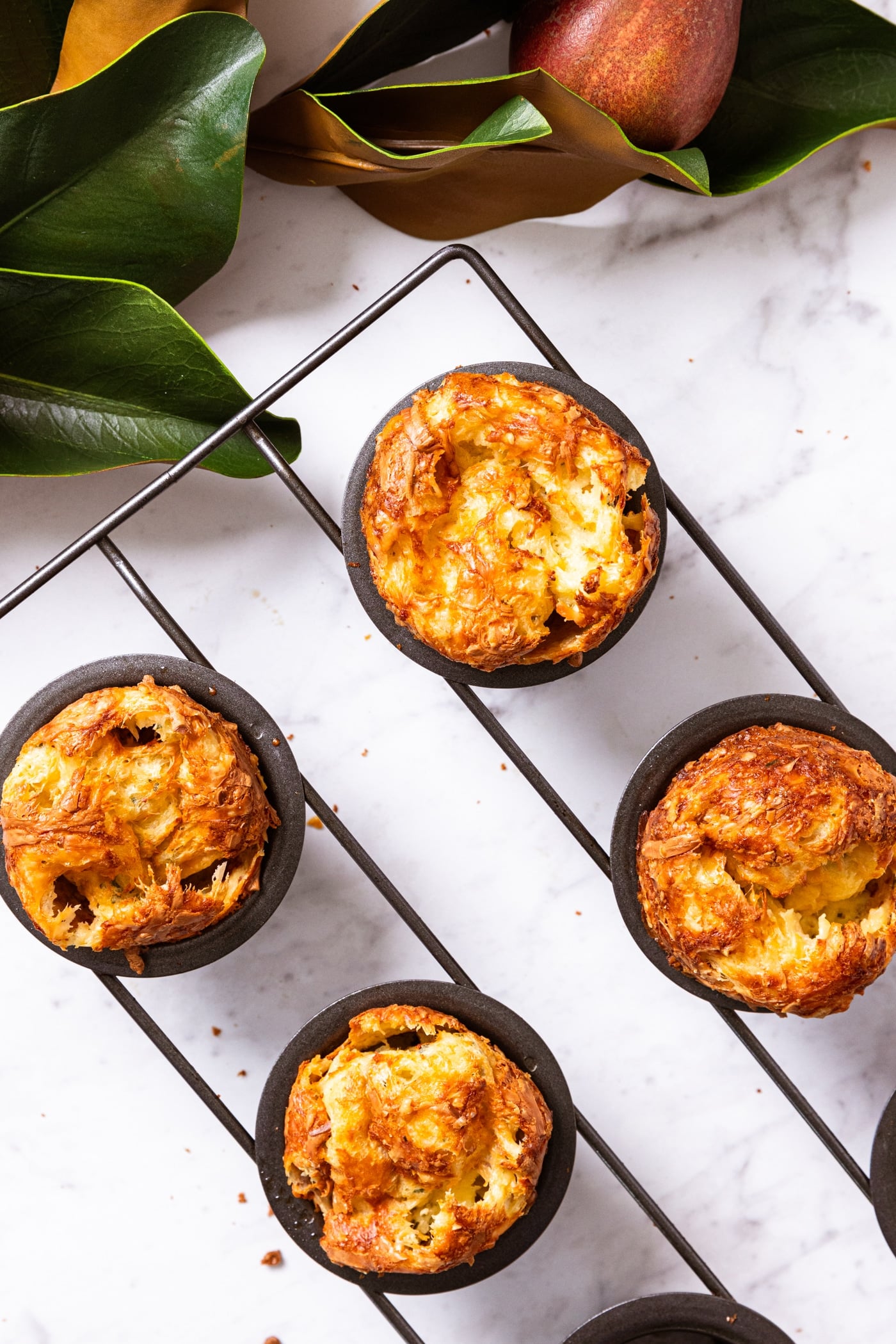 Marble surface with magnolia garland and pears along the top with black popover pan with baked and golden popovers still sitting in pan.